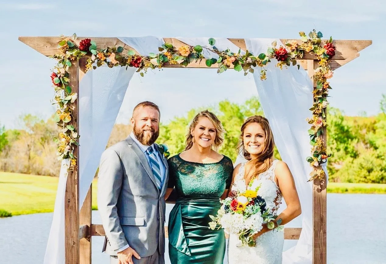 A bride with long brown hair holding a bouquet of flowers, a man in a gray suit, and a woman in a green dress standing under a decorated wedding arch near a body of water with trees in the background.