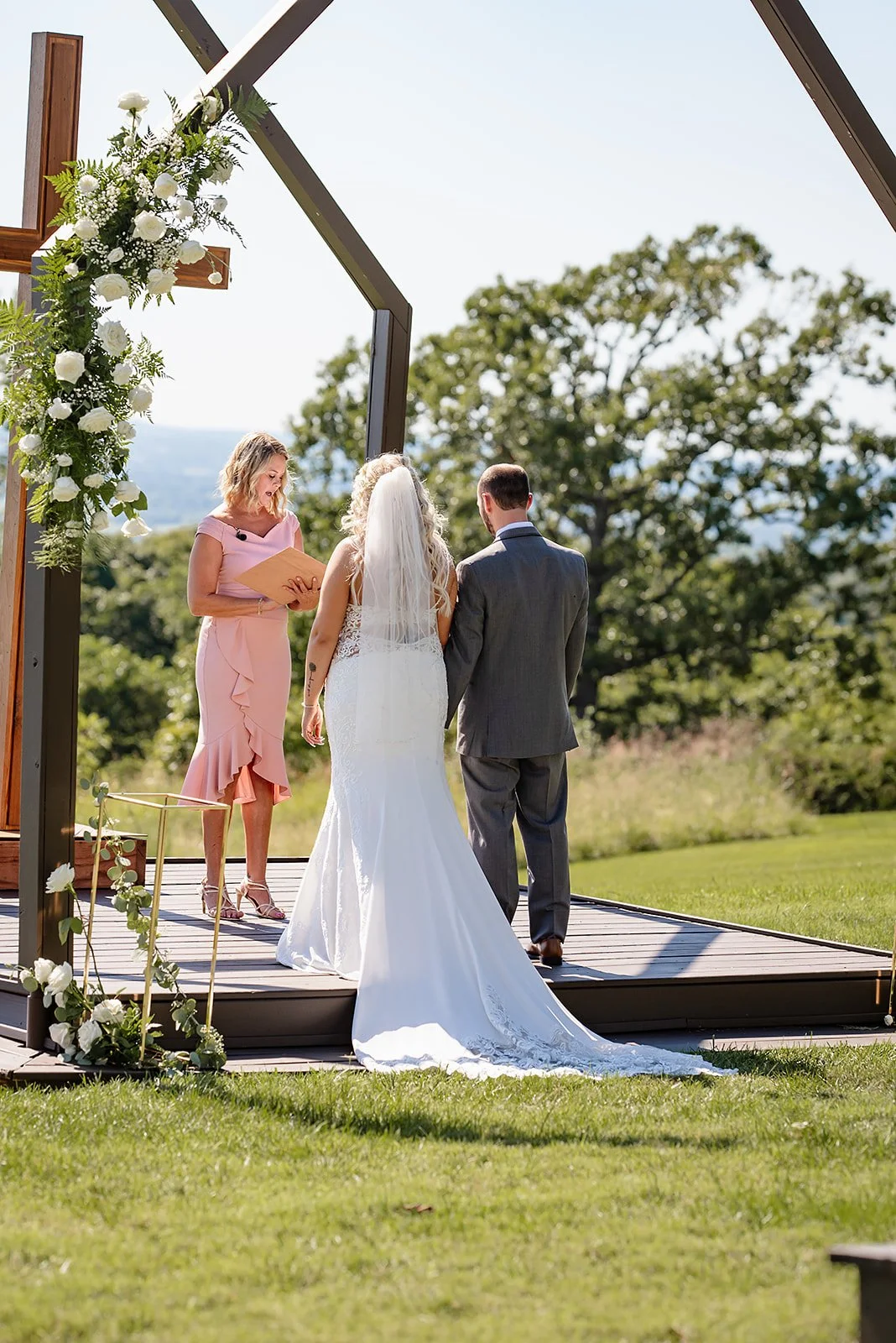 A couple getting married outdoors on a wooden platform decorated with white flowers, with an officiant in a pink dress officiating the ceremony, and trees and greenery in the background.