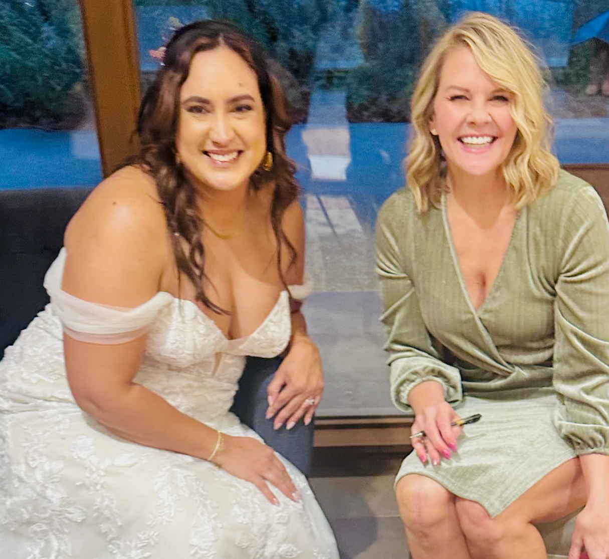 Two women sitting next to each other, smiling at the camera, inside a room with large windows and a scenic outdoor view behind them, one in a white dress and the other in a green dress.