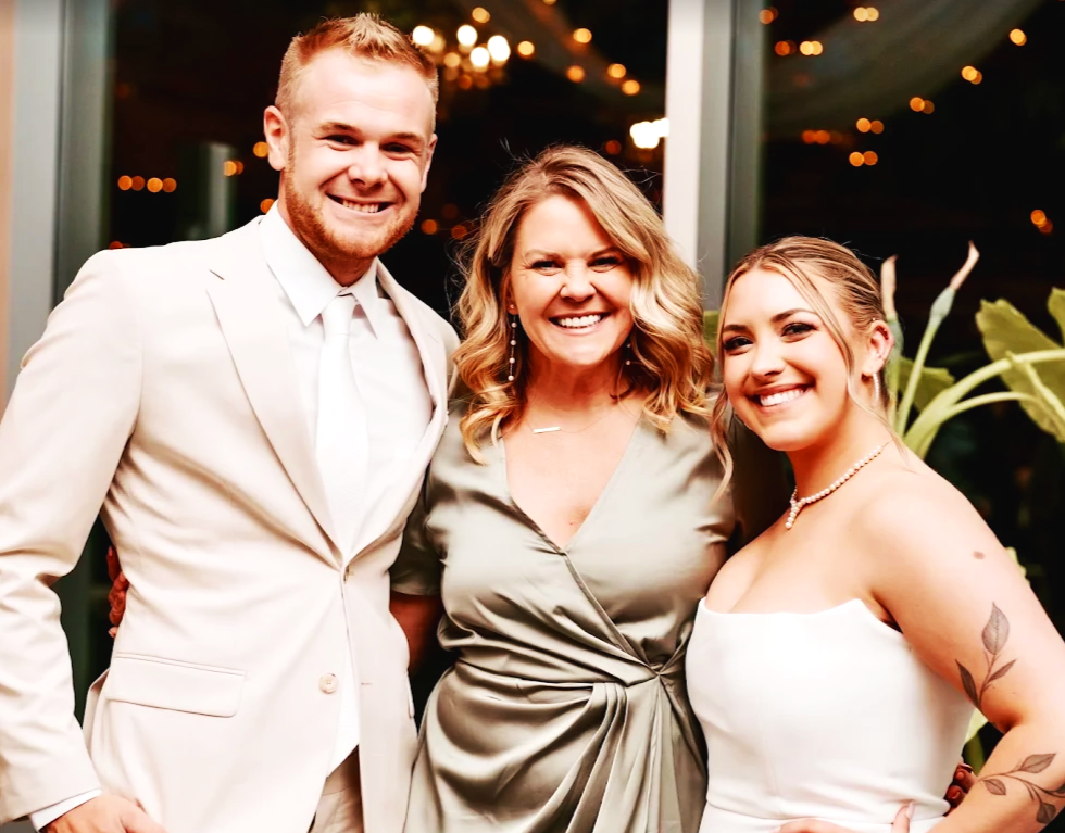 Three people dressed in formal attire smiling at an indoor event at night, with string lights visible through the window behind them.