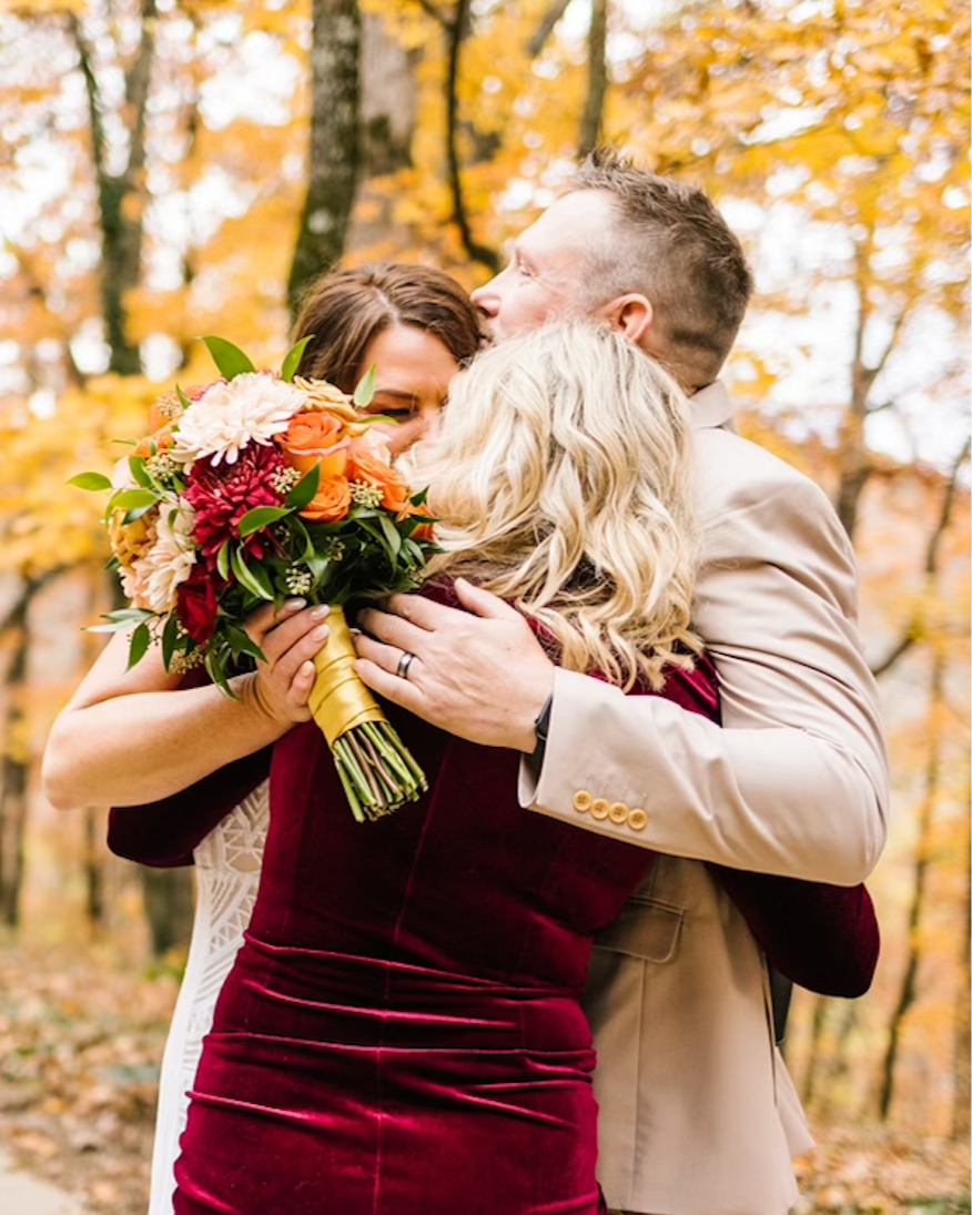 Four people hugging outdoors during autumn, with trees in fall foliage in the background. One woman is holding a bouquet of orange, red, and white flowers.