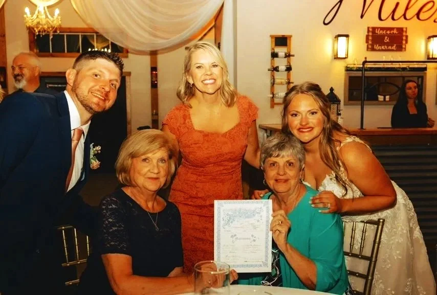 A group of six people at a celebration, with three women sitting and two women and one man standing behind them, holding a certificate.