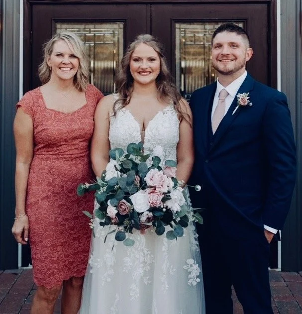 Three people standing in front of a wooden door, smiling. The woman in the center is wearing a white wedding dress and holding a bouquet of pink and white flowers with green foliage. The woman on the left is wearing a pink lace dress, and the man on 