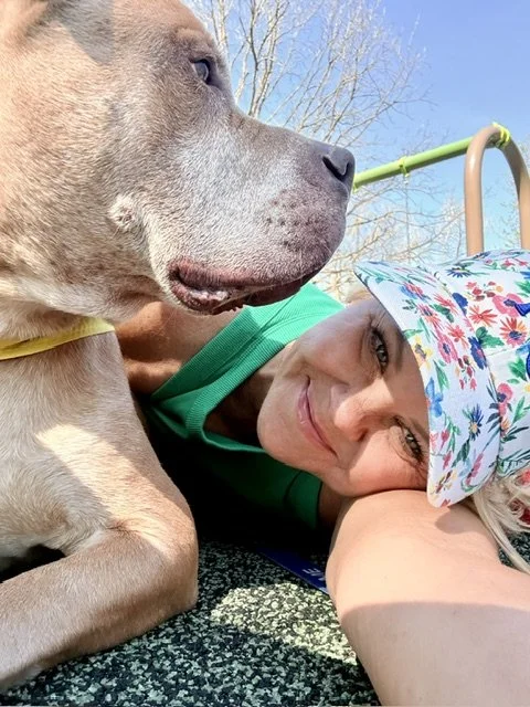 A woman in a colorful hat and green shirt lying on the ground next to a large dog with a tan coat. The woman is smiling and resting her head on her arm, with a playground in the background and clear blue sky.