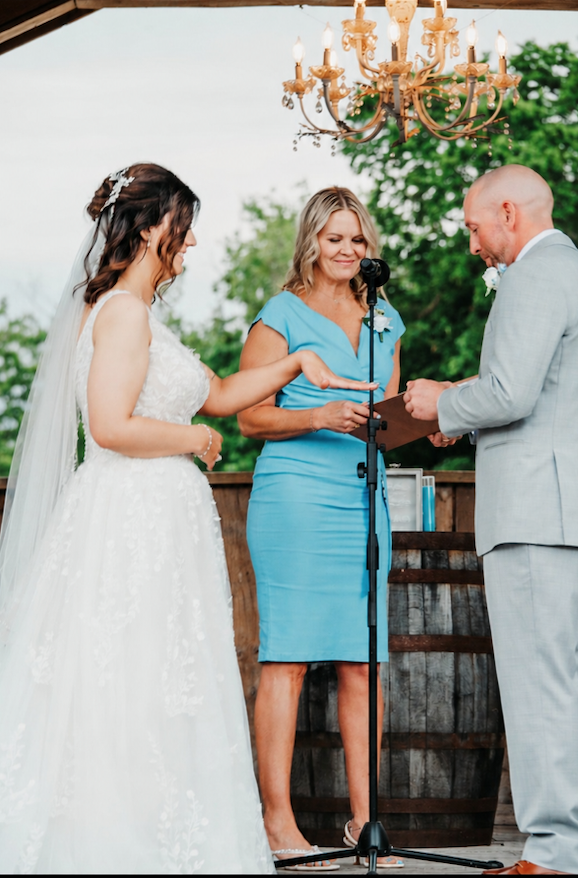 A bride and groom exchanging vows during their outdoor wedding ceremony, with a woman officiant, under a chandelier, with trees in the background.