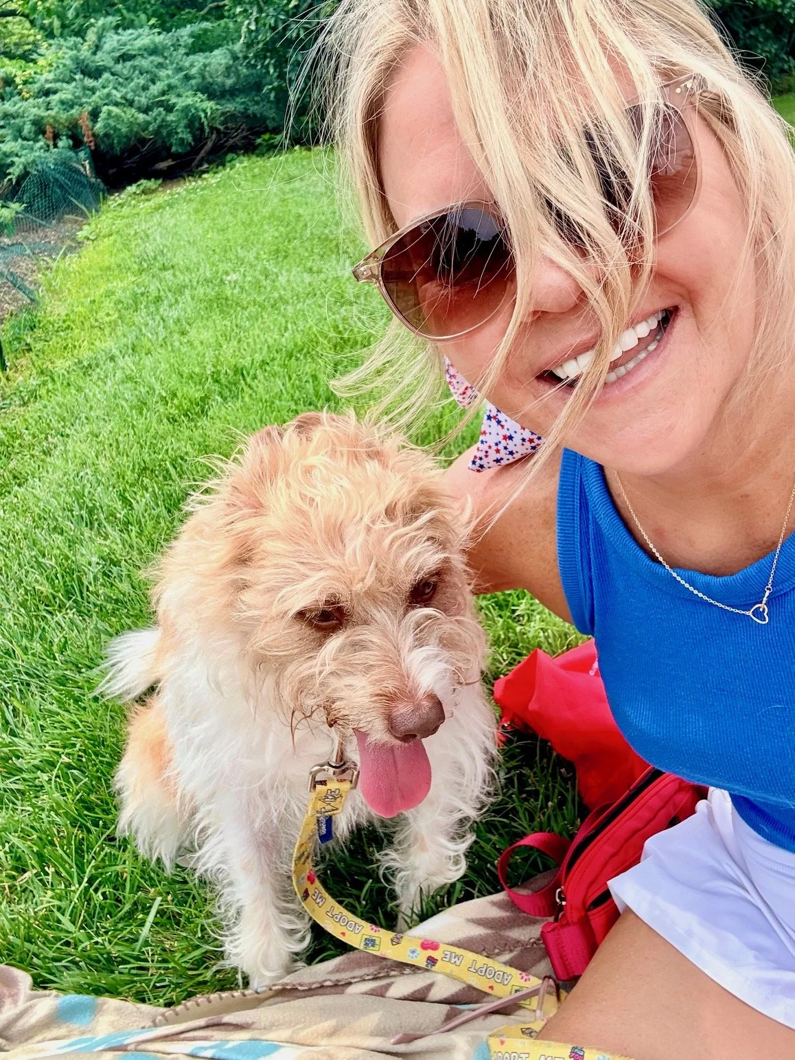 A woman with blonde hair, wearing sunglasses and a blue shirt, smiling while sitting on green grass with a beige and white dog. The dog has its tongue out and a yellow leash. They are outdoors in a grassy area with trees in the background.