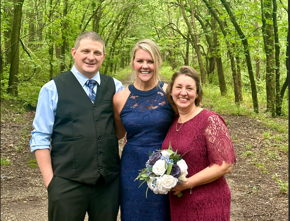 Three people standing outdoors in a wooded area, dressed formally and smiling at the camera. One woman is holding a bouquet of white and purple flowers.
