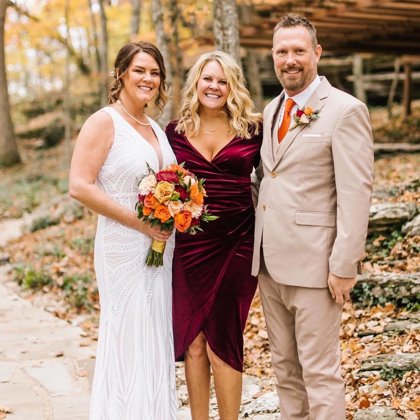 A smiling woman in a white dress holding a bouquet of orange, pink, and yellow flowers, standing outdoors with two smiling people, a woman in a burgundy dress and a man in a beige suit with an orange tie, during an autumn wedding in a wooded area wit