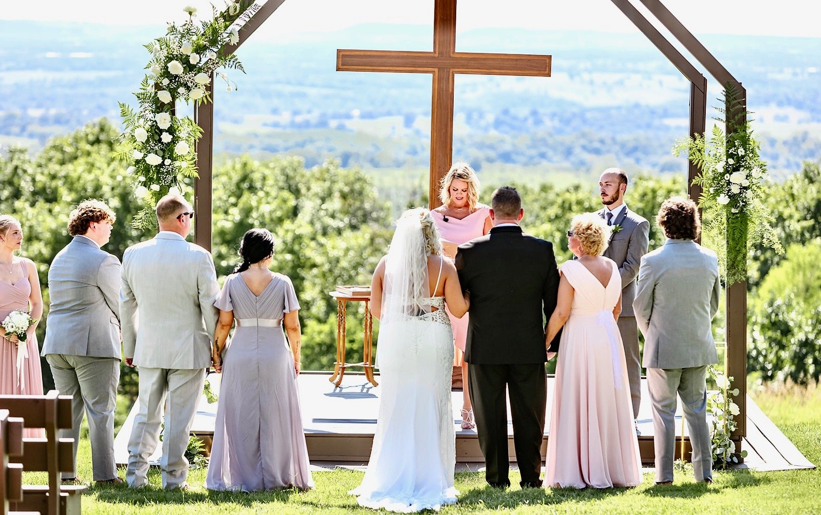 Outdoor wedding ceremony with a bride and groom facing an officiant, surrounded by wedding party members, under a decorated arch with a large wooden cross, set against a green landscape background.