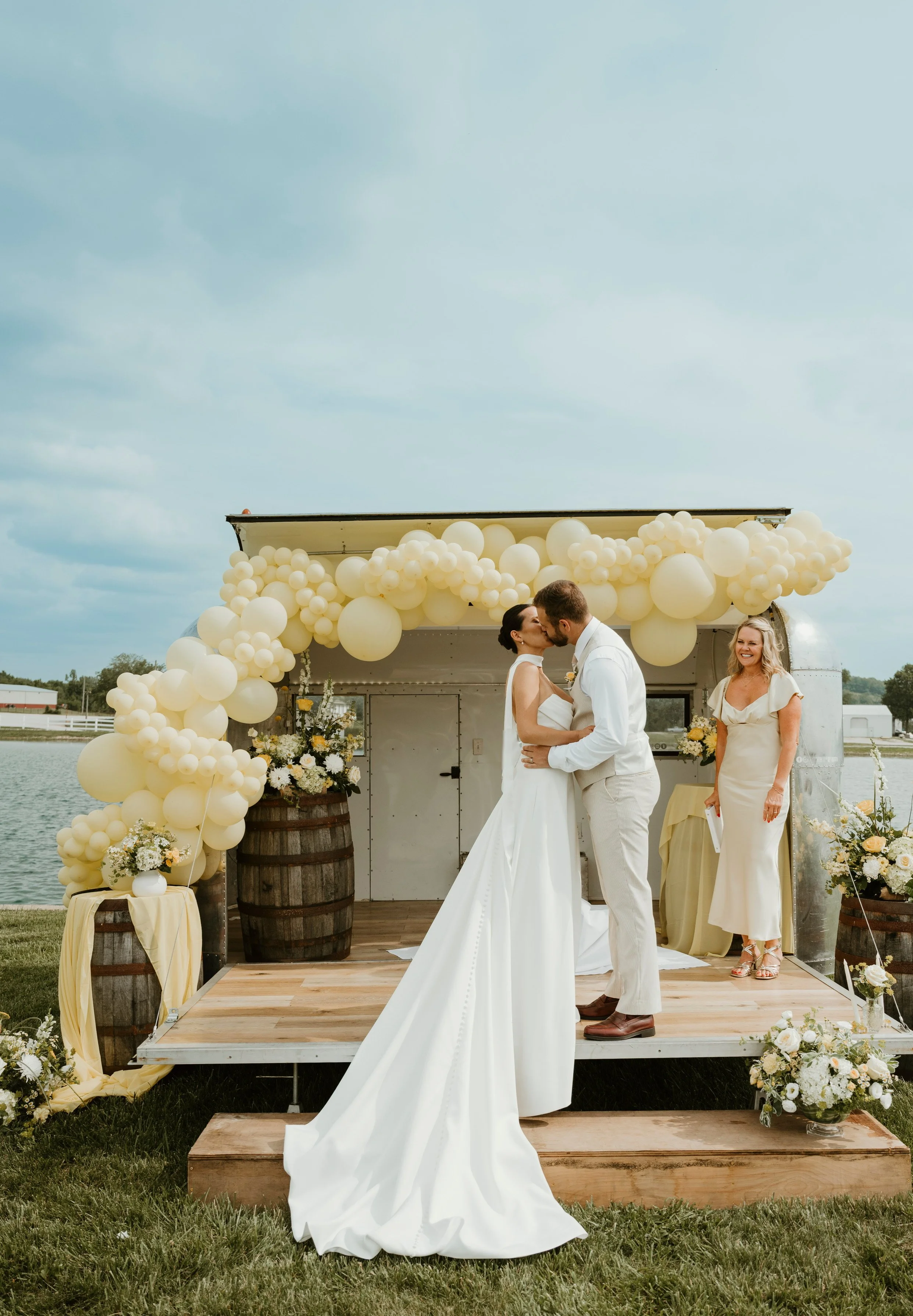 A couple shares a kiss during their outdoor wedding ceremony on a small wooden stage decorated with cream-colored balloons and flowers, with a woman in a beige dress smiling nearby.
