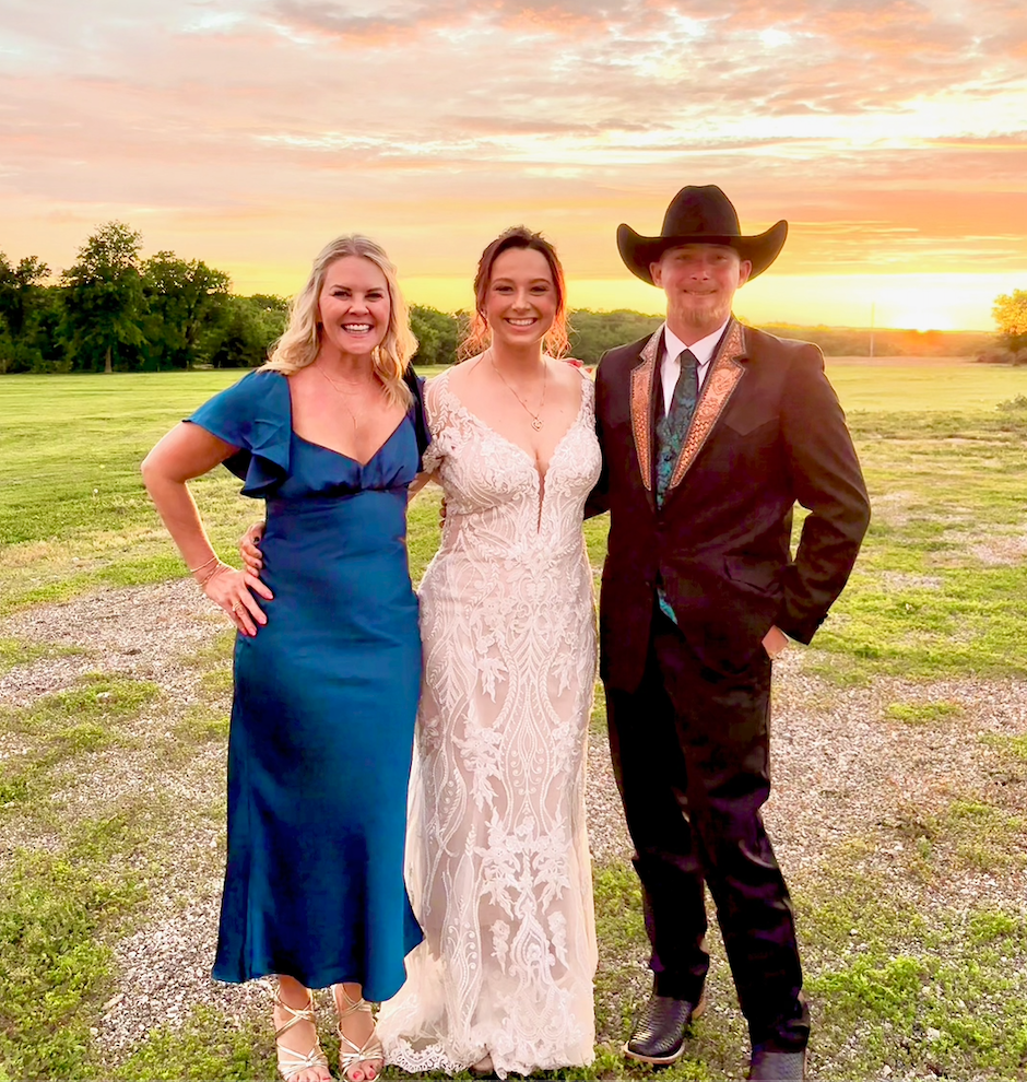 Three people standing together outdoors during sunset. On the left, a woman in a blue dress with ruffled sleeves, the middle a woman in a white wedding dress, and on the right a man in a black suit with a large black cowboy hat, smiling.