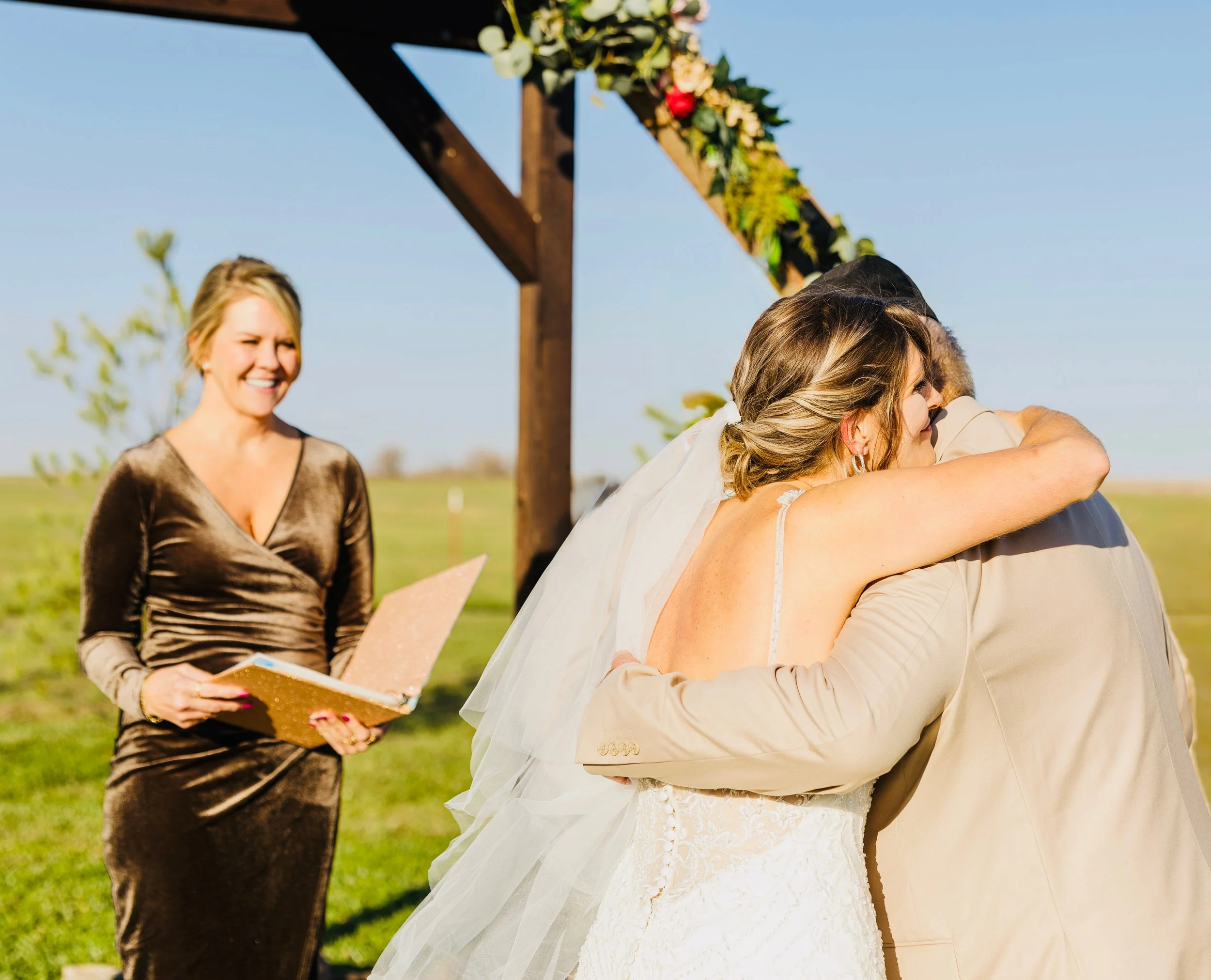 A wedding ceremony outdoors with a bride and groom embracing, an officiant smiling and holding a book, under a wooden arch decorated with flowers and greenery, with a clear blue sky in the background.