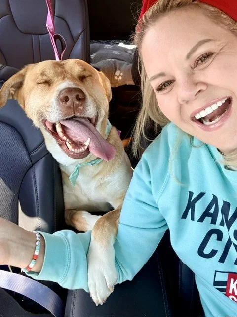 Woman smiling with a red headband in her hair and a commercial t-shirt, taking a selfie with a happy, tan and white dog in a car, the dog is sitting in a car seat with eyes closed and tongue out.