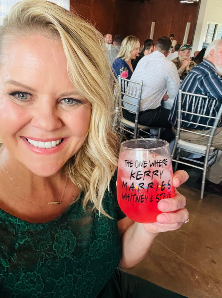 A smiling woman with blonde hair holding a glass of red beverage at a wedding reception. The glass has a message written on it: "The one where Kerry marries Whitney & Steve." There are multiple people seated at tables in the background.