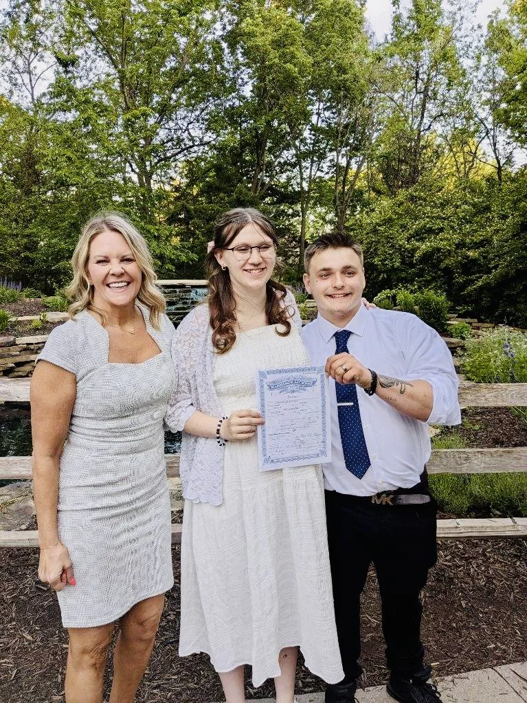 Three people standing outdoors in front of trees, smiling and posing for a photo. The woman on the left is wearing a light gray dress, the woman in the middle is wearing a white dress and glasses, holding a document, and the man on the right is weari