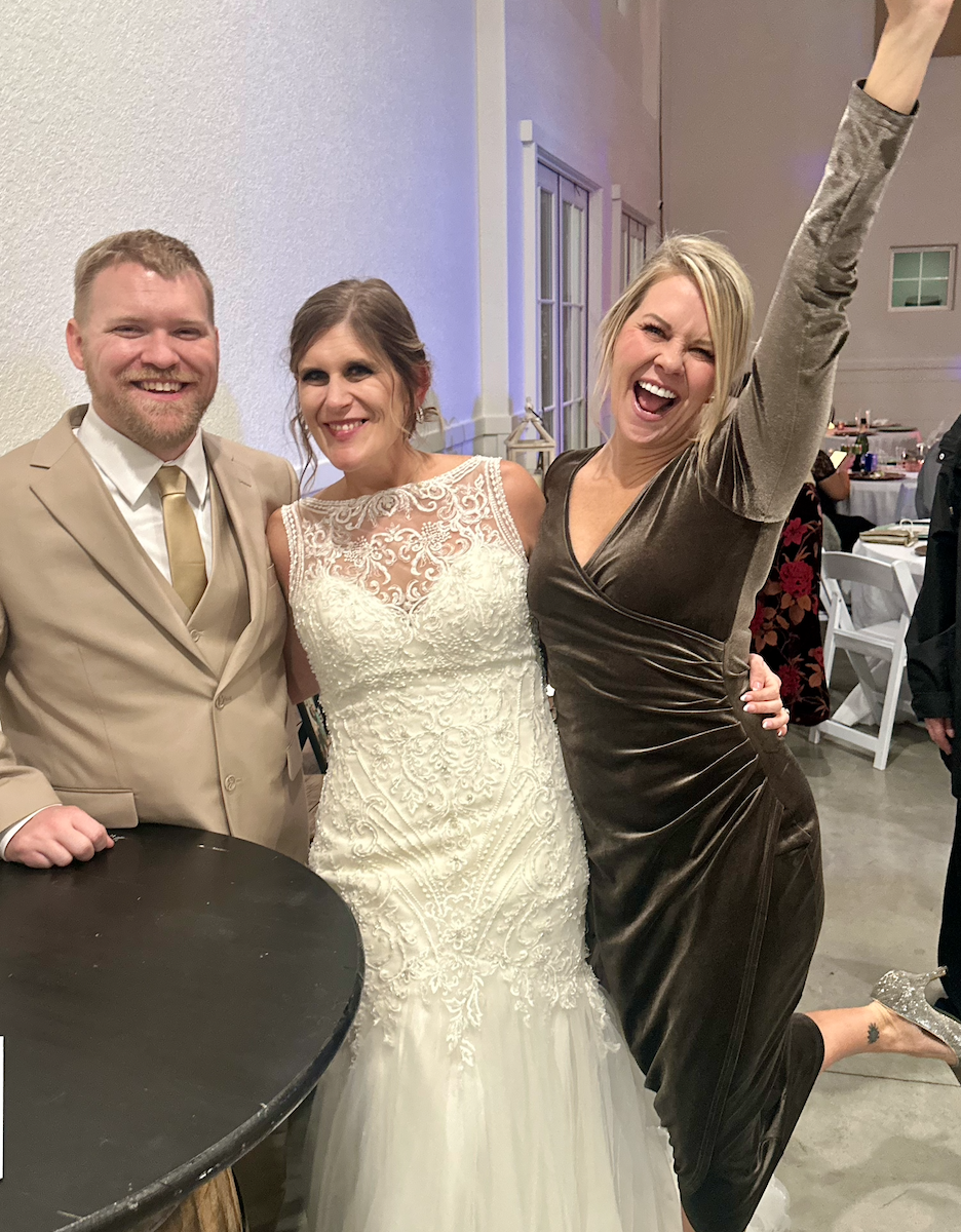 Three people at a celebration, two women and a man, all smiling and posing for the photo. The woman in the middle is wearing a white lace wedding dress. The man on the left is wearing a beige suit with a gold tie. The woman on the right is wearing a 