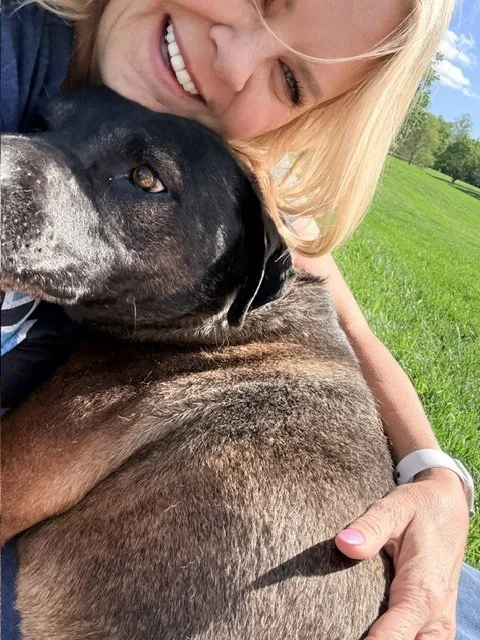 A woman with blonde hair smiling and hugging a black and white dog outdoors on a sunny day with green grass and trees in the background.