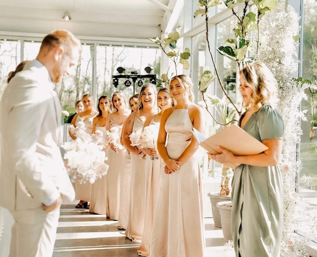 A bride and a groom standing in front of a wedding party of bridesmaids during a wedding ceremony in a bright, sunlit indoor space surrounded by green plants.