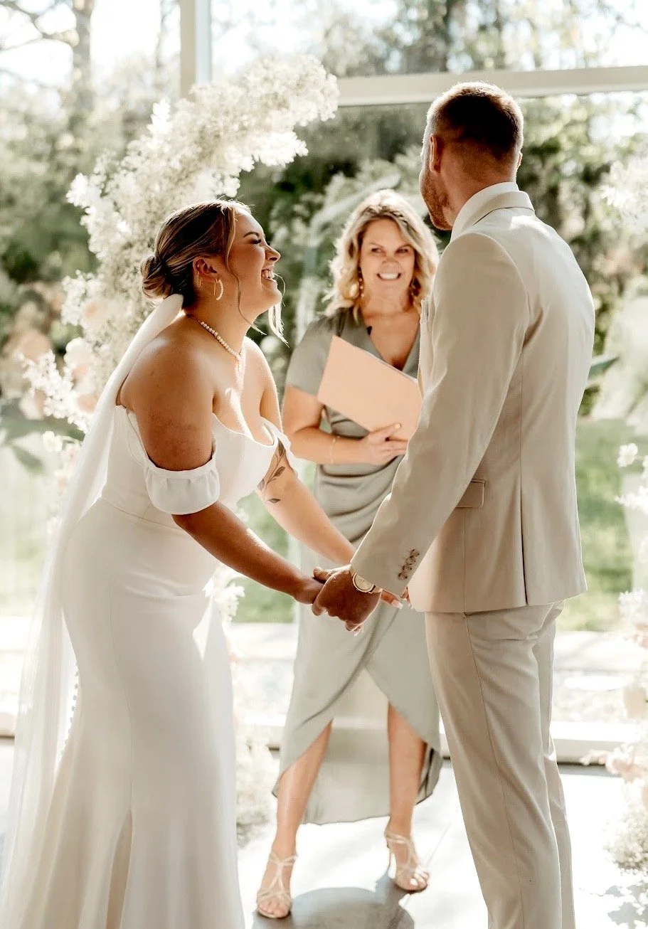 Bride and groom holding hands during their wedding ceremony, smiling at each other, with an officiant standing behind them, in a bright, indoor venue with large windows and floral decorations.