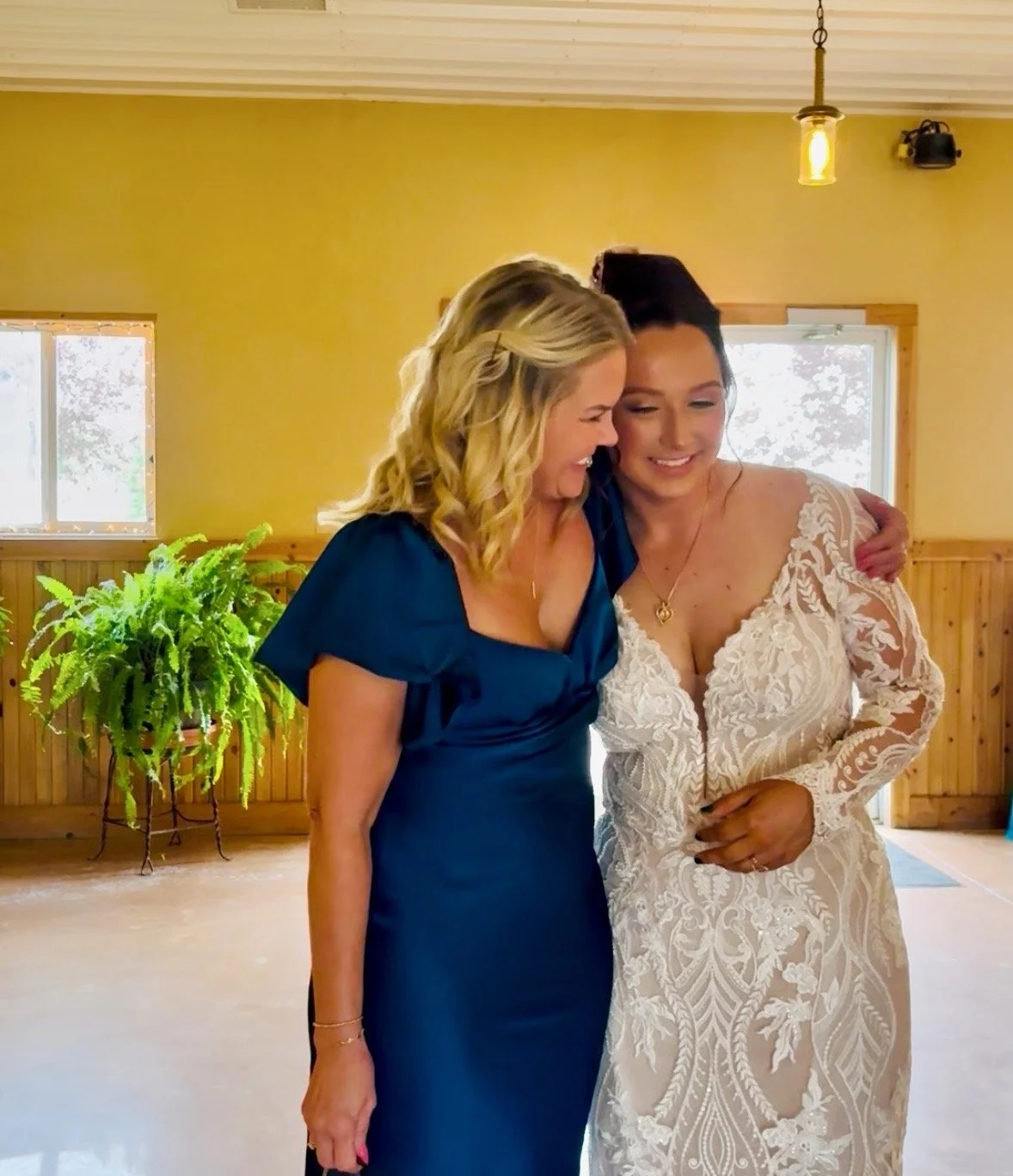 Two women smiling and hugging in a warmly lit room with wooden walls and two windows. One woman is wearing a white lace dress and the other a blue dress.
