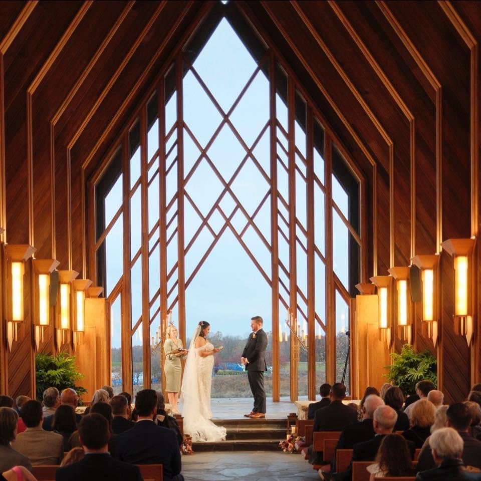 A couple exchanges vows during a wedding ceremony inside a modern wooden chapel, with guests seated and a scenic outdoor view in the background.