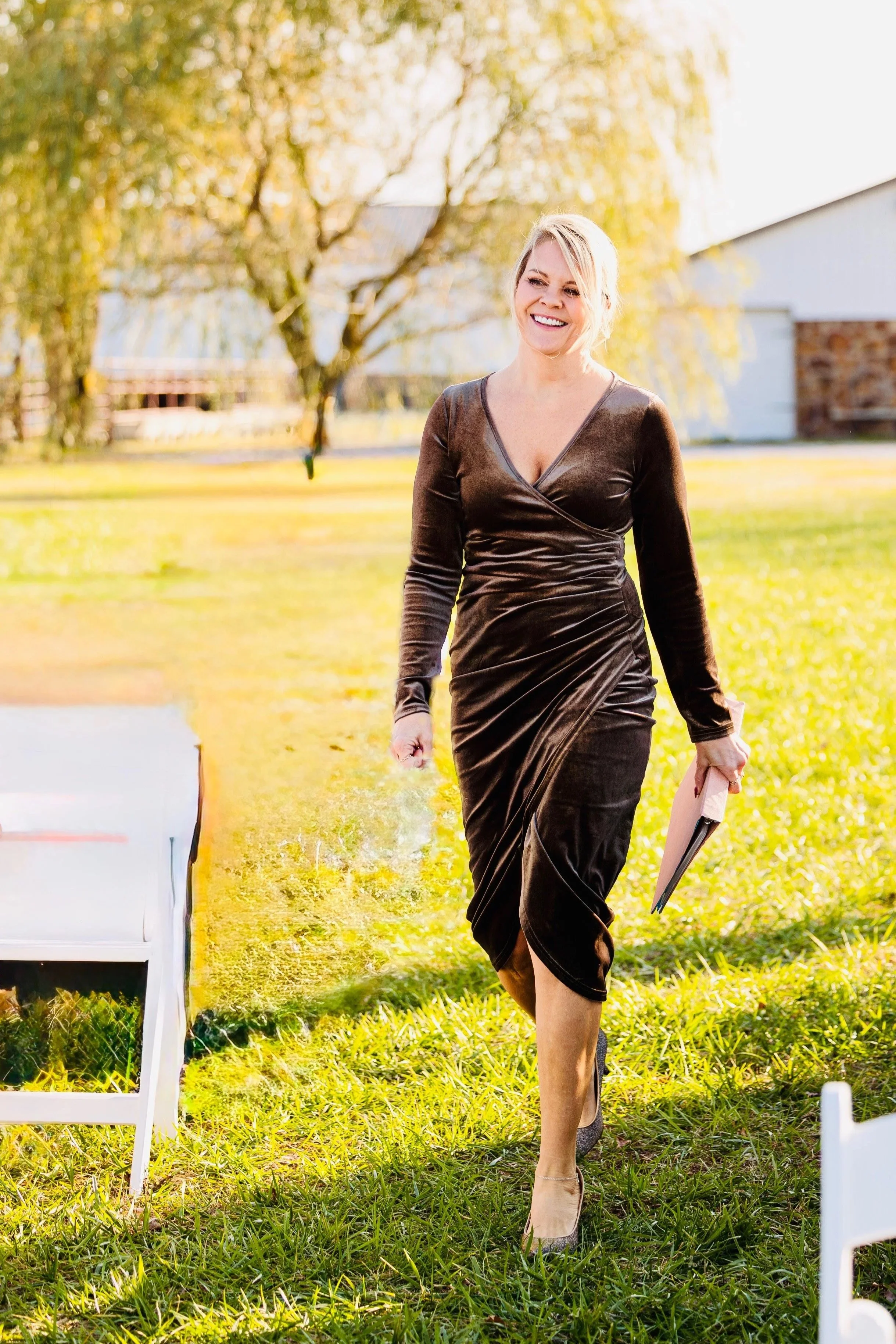 A woman walking outdoors in a black velvet dress holding a clutch bag, smiling, with trees and a building in the background, sunny weather.