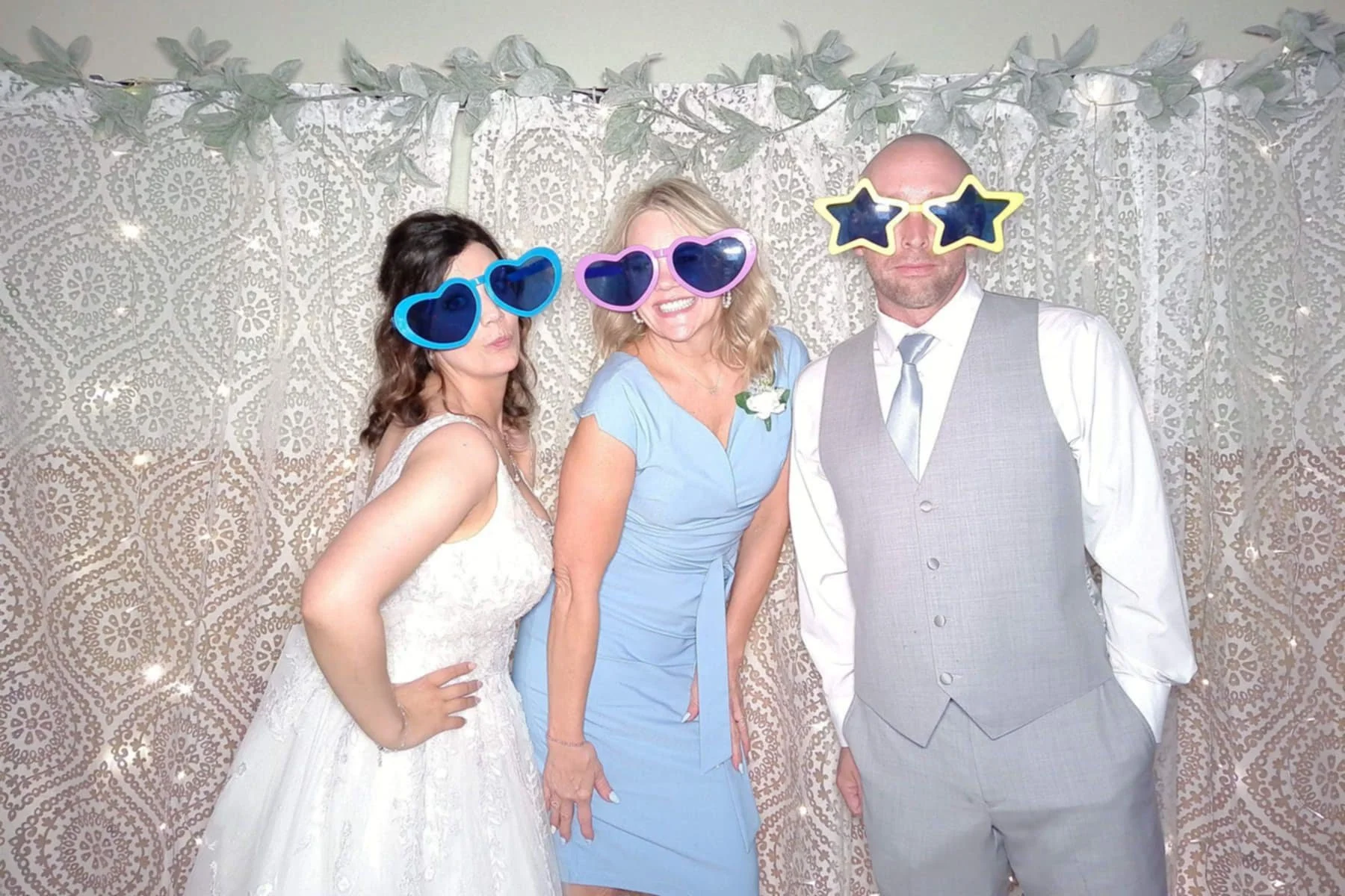 Three people at a celebration, wearing colorful oversized sunglasses with a decorative patterned backdrop. The woman in the middle is smiling, dressed in a light blue dress with a boutonniere, and the man on the right is dressed in a gray vest and wh