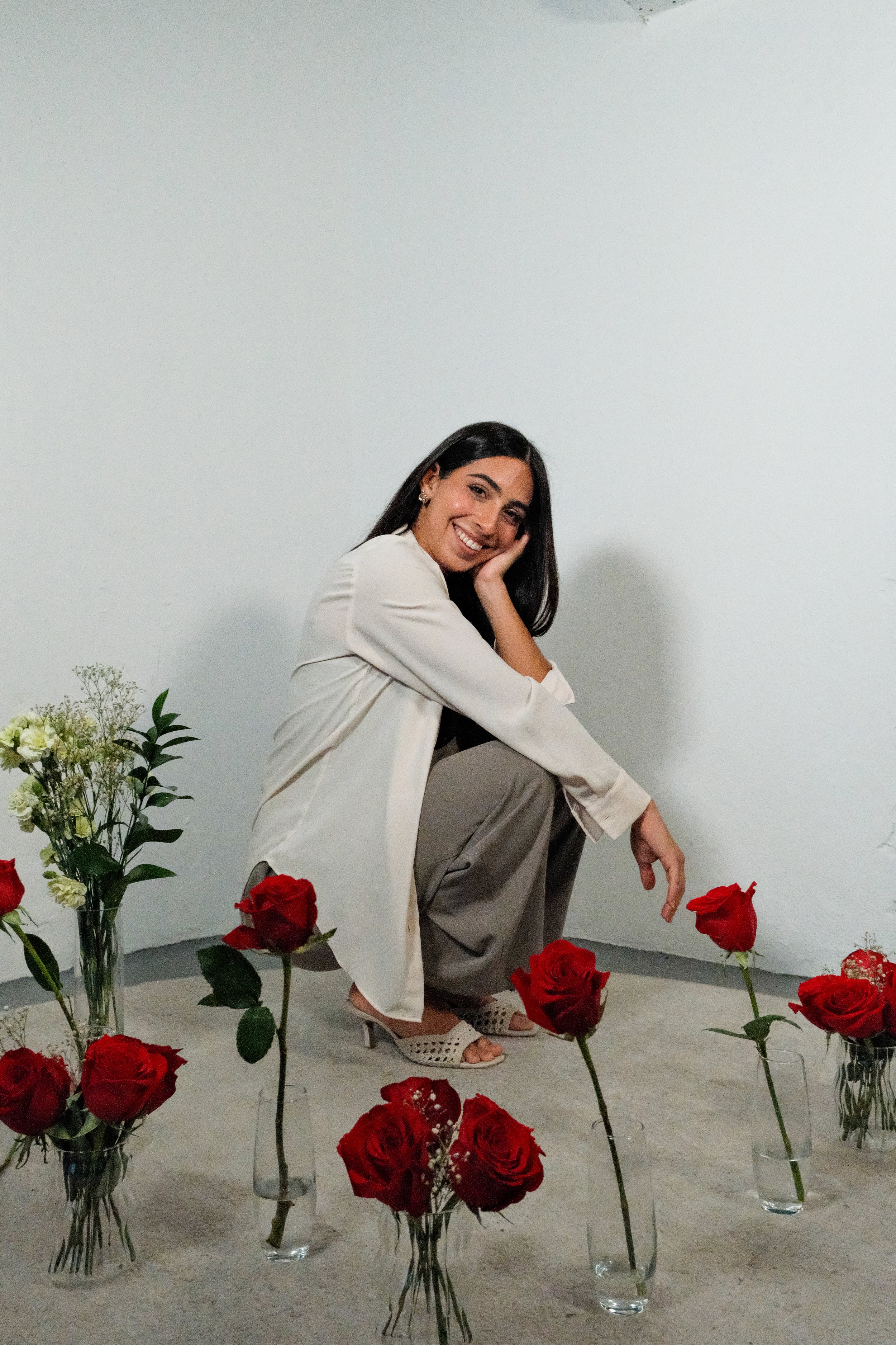 A smiling woman with long dark hair, sitting among red roses in glass vases, wearing a cream-colored blazer, beige pants, and open-toed high heels, posing with a relaxed, cheerful demeanor against a plain white wall.