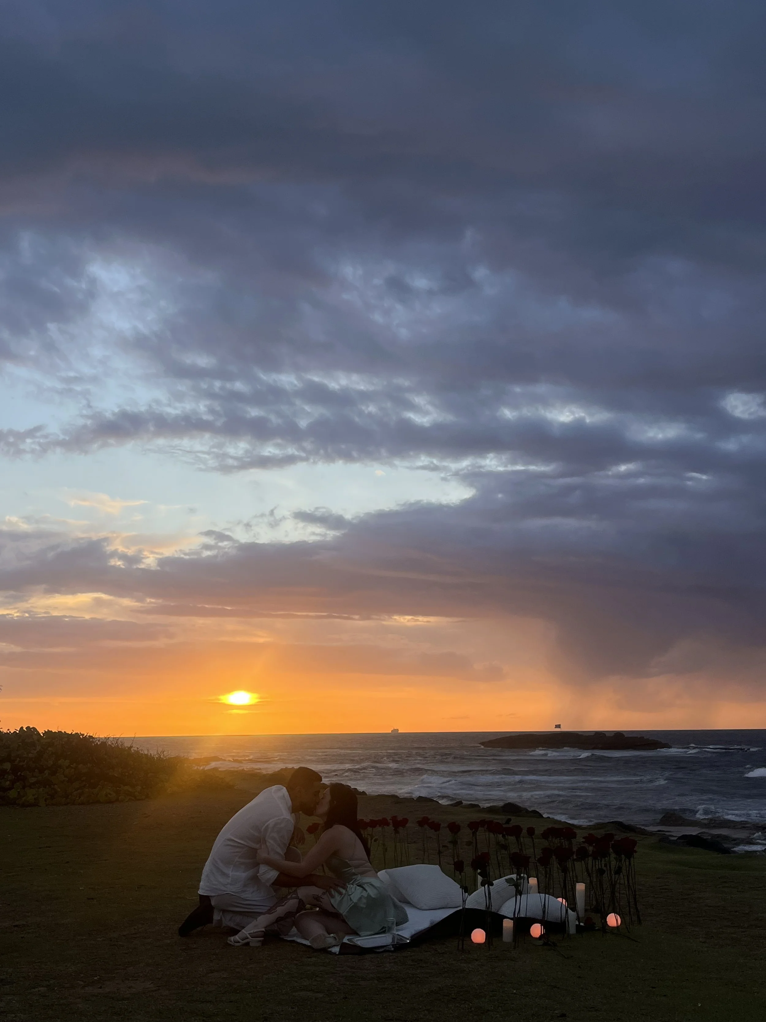 A couple sitting on a blanket by the water during sunset, with candles and flowers around them.