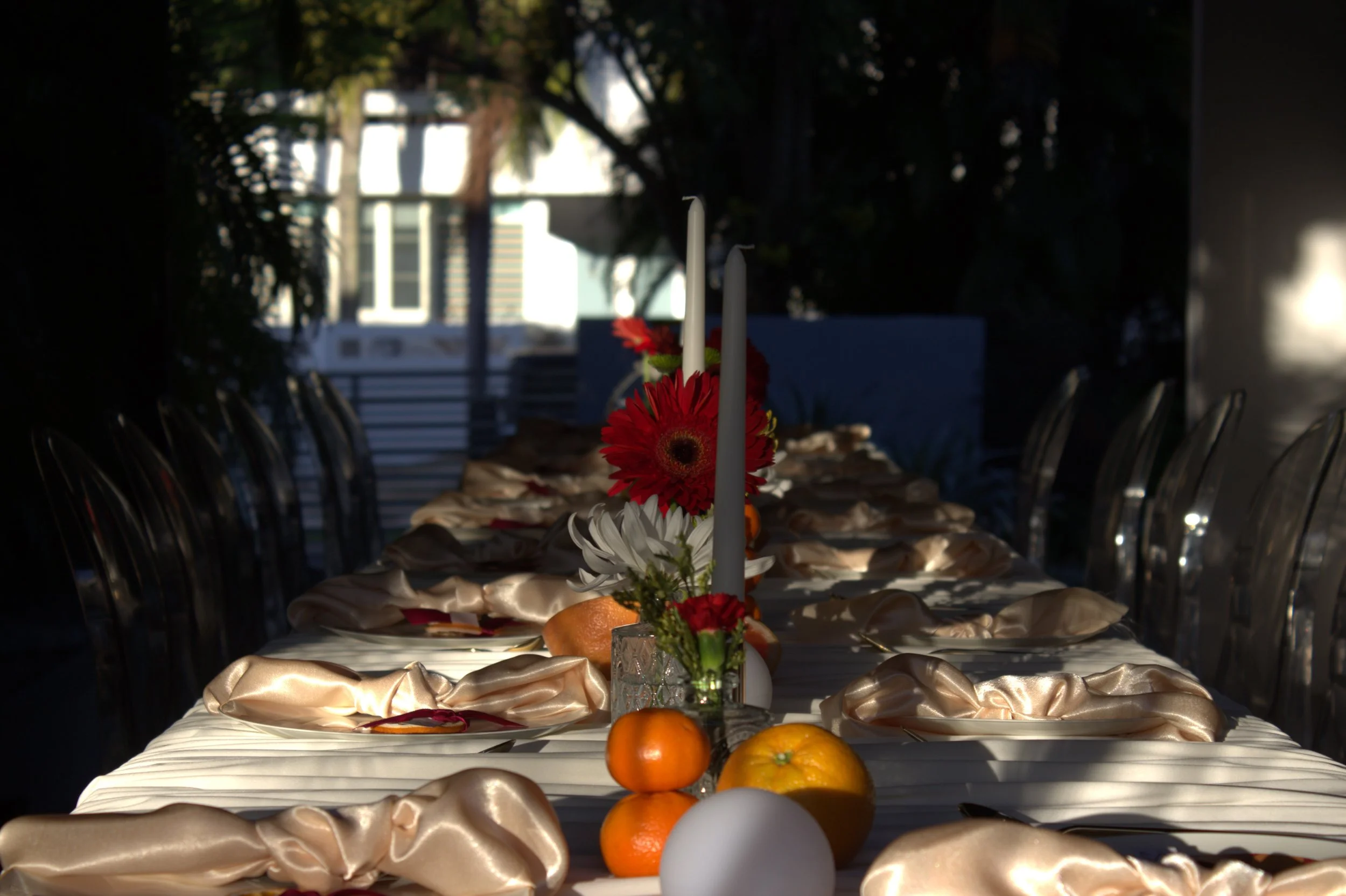 A long dining table with a satin tablecloth, decorated with a floral centerpiece and various fruits, set outdoors under trees with a building in the background.