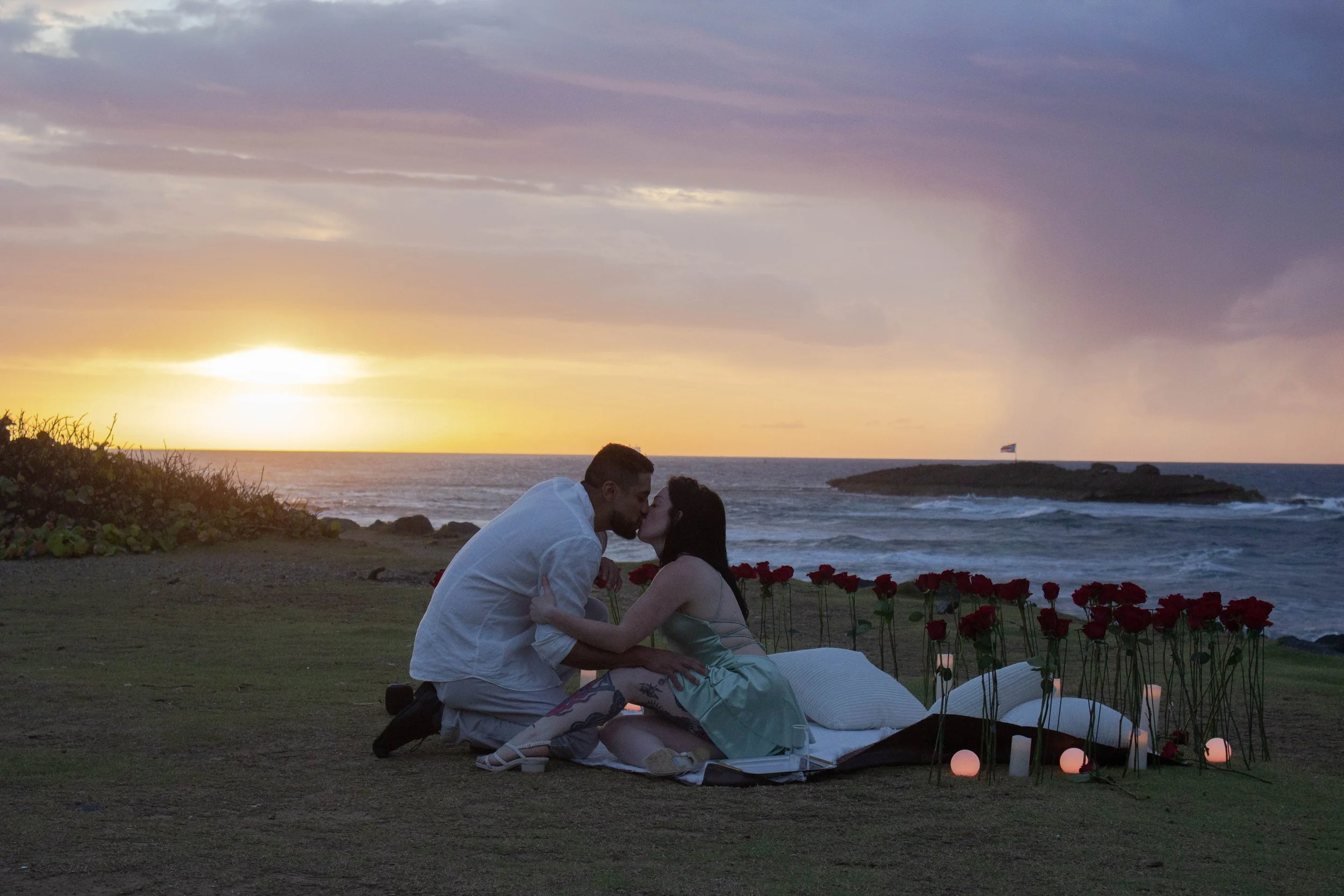 A couple sharing a kiss during a romantic sunset on a beach with a small boat, red roses, and candles nearby.