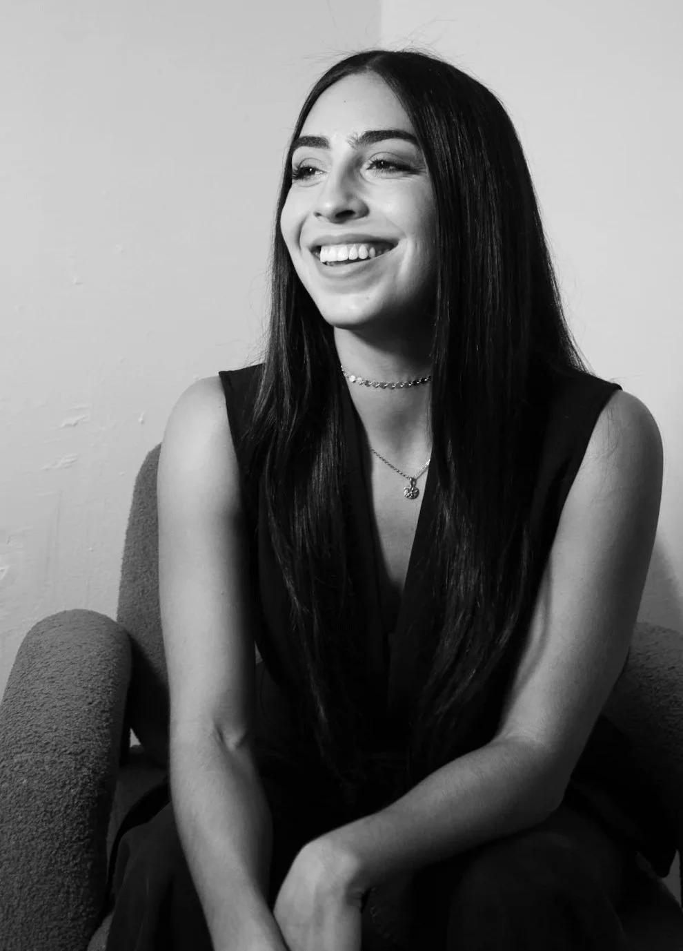 Black and white photo of a smiling woman with long dark hair, wearing a sleeveless top, a choker necklace, and a pendant necklace, sitting on a textured armchair against a plain wall background.