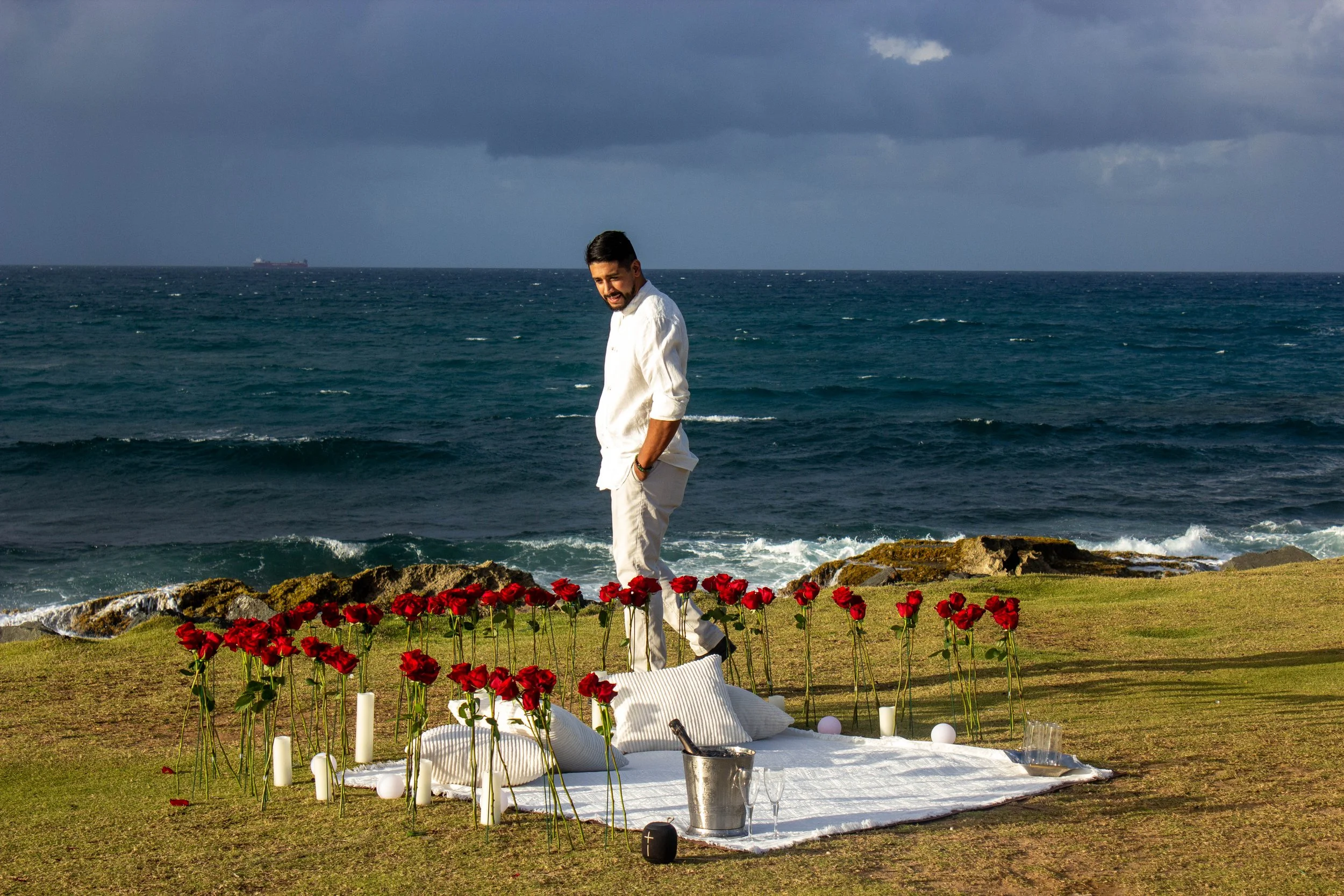 A man dressed in white standing by a memorial with red roses, candles, and a white cloth near the ocean, under a cloudy sky.