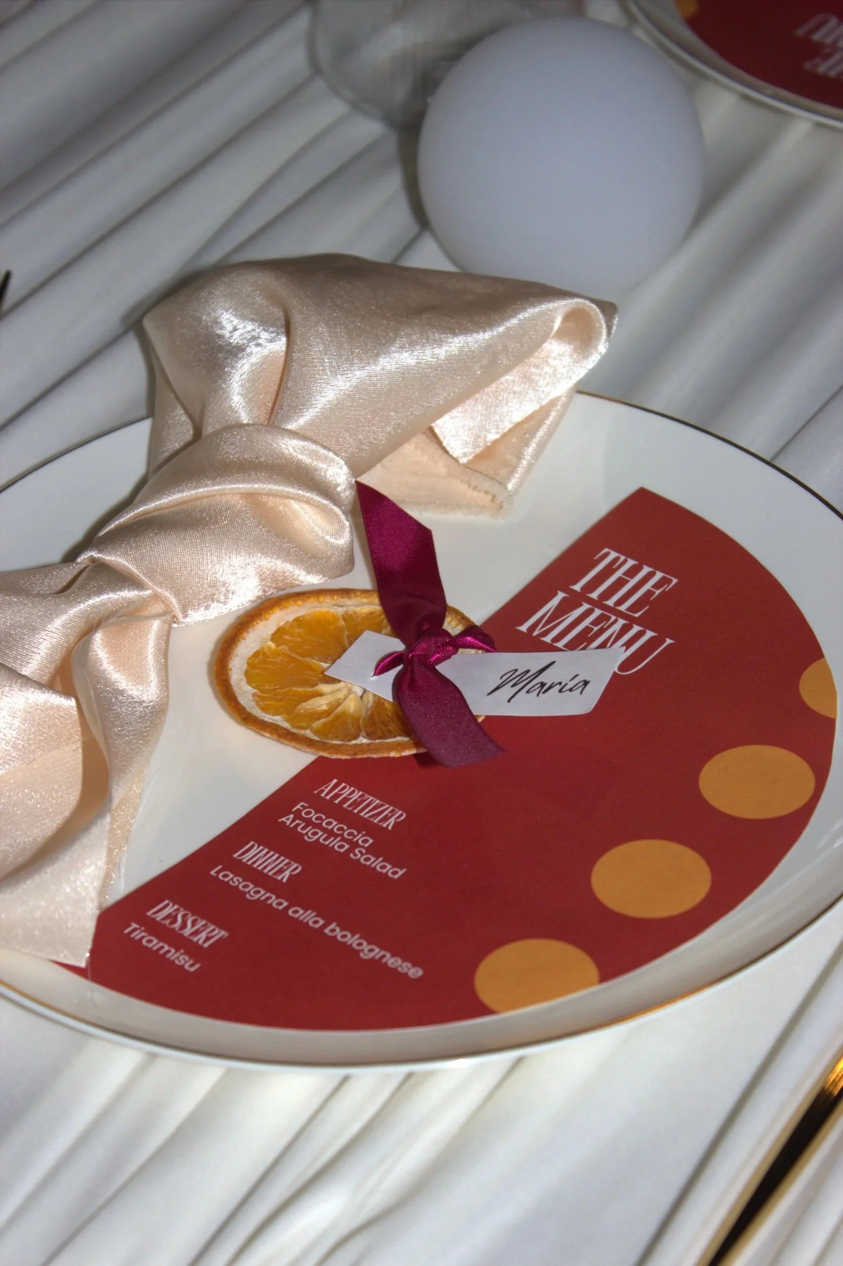 A menu on a red background with yellow dots, placed on a white surface. A satin beige bow and a slice of dried orange are also visible on the menu.