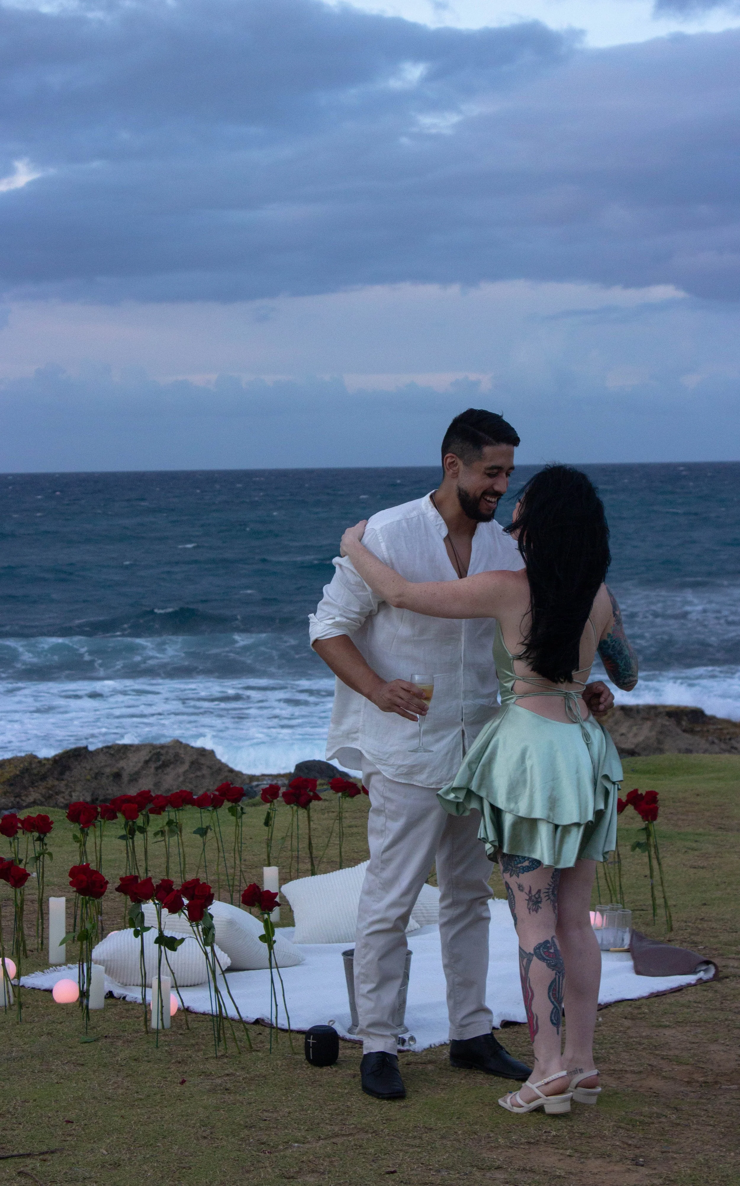 A couple is standing close together on a grassy area near the ocean, smiling and looking into each other's eyes during a romantic outdoor event with a decorated setup of flowers and candles on the ground.