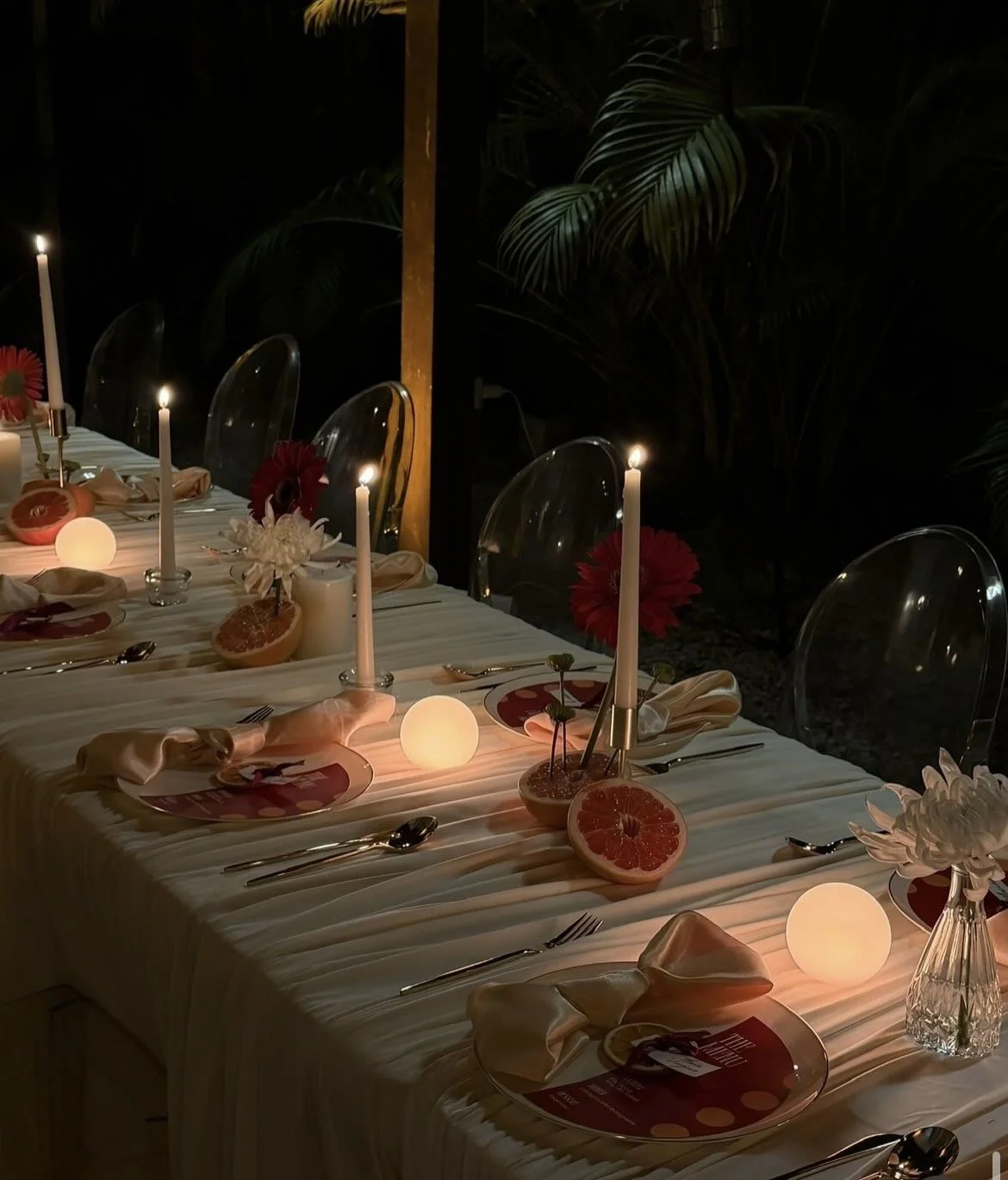 A long dining table set for an outdoor dinner at night, decorated with candles, sliced grapefruits, flowers, and place settings with napkins and utensils.