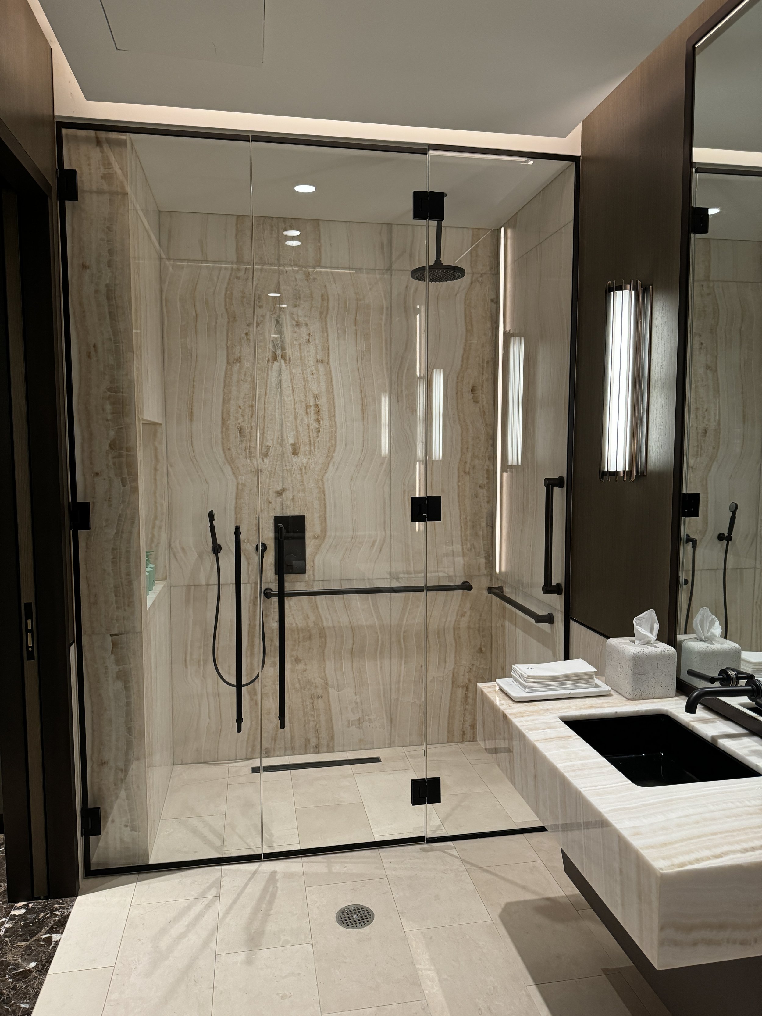 Modern bathroom with a glass-enclosed shower, marble walls, a marble vanity with a black sink, and a mirror with vertical lighting.