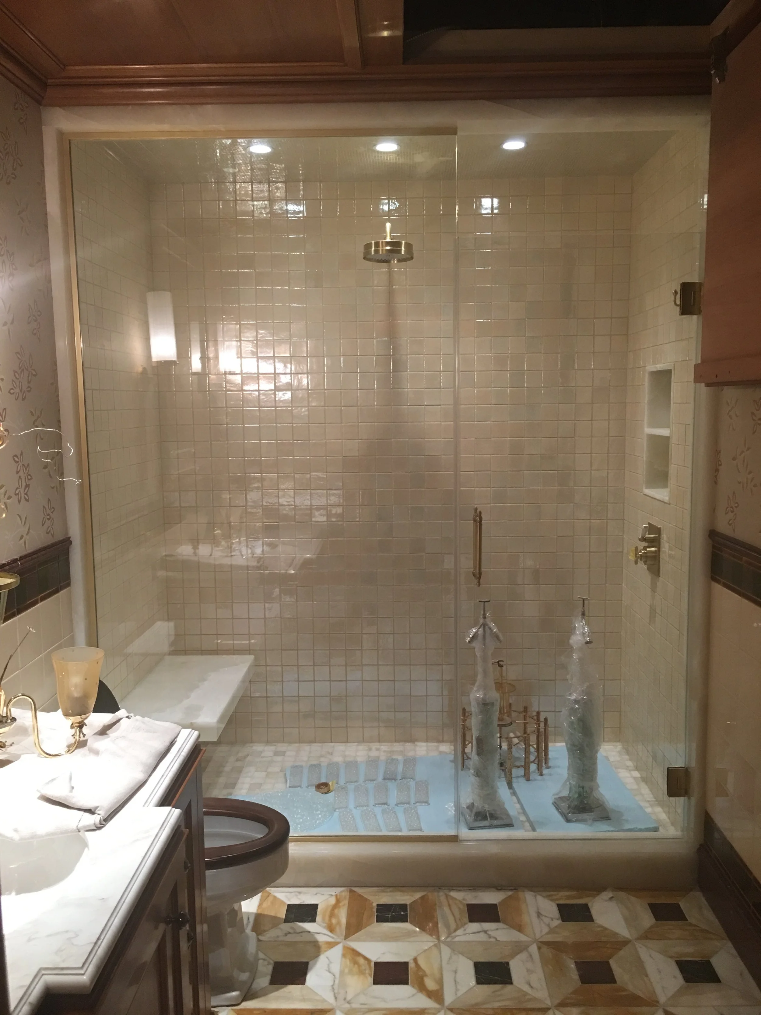 Bathroom with beige tiled shower area, glass door, marble countertop with a soap dish and mirror, patterned tile flooring, and unopened plumbing fixtures.