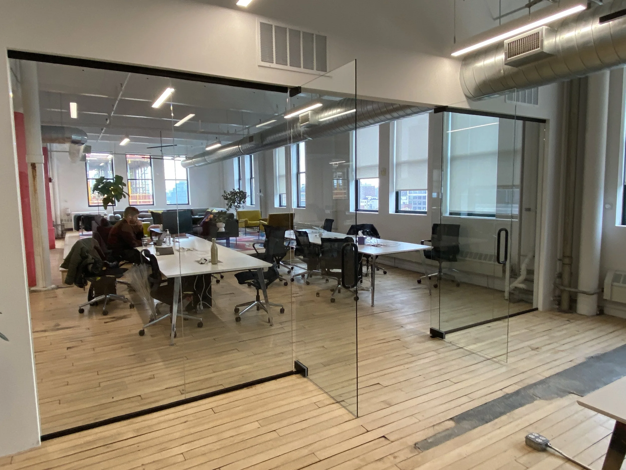A modern office space seen through a glass partition, with wooden flooring, tables, chairs, and large windows showing a cityscape outside.