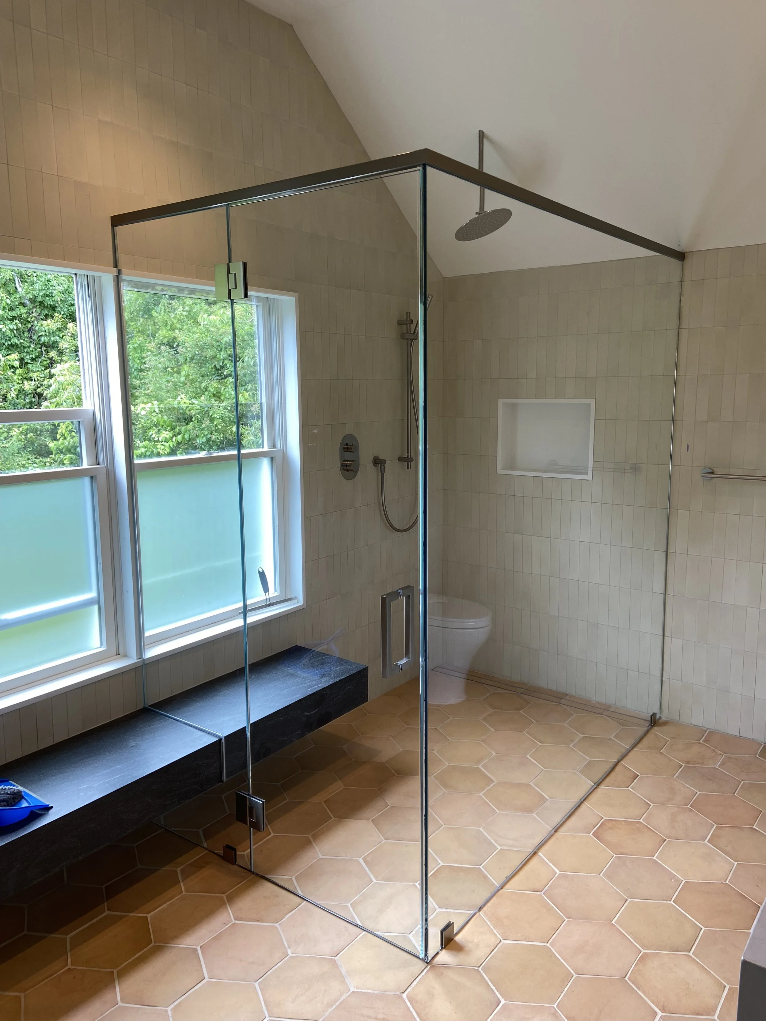 A bathroom with a walk-in glass shower enclosure. The shower has a rain shower head and a handheld shower. There is a built-in niche in the wall. A window with a view of green trees provides natural light.
