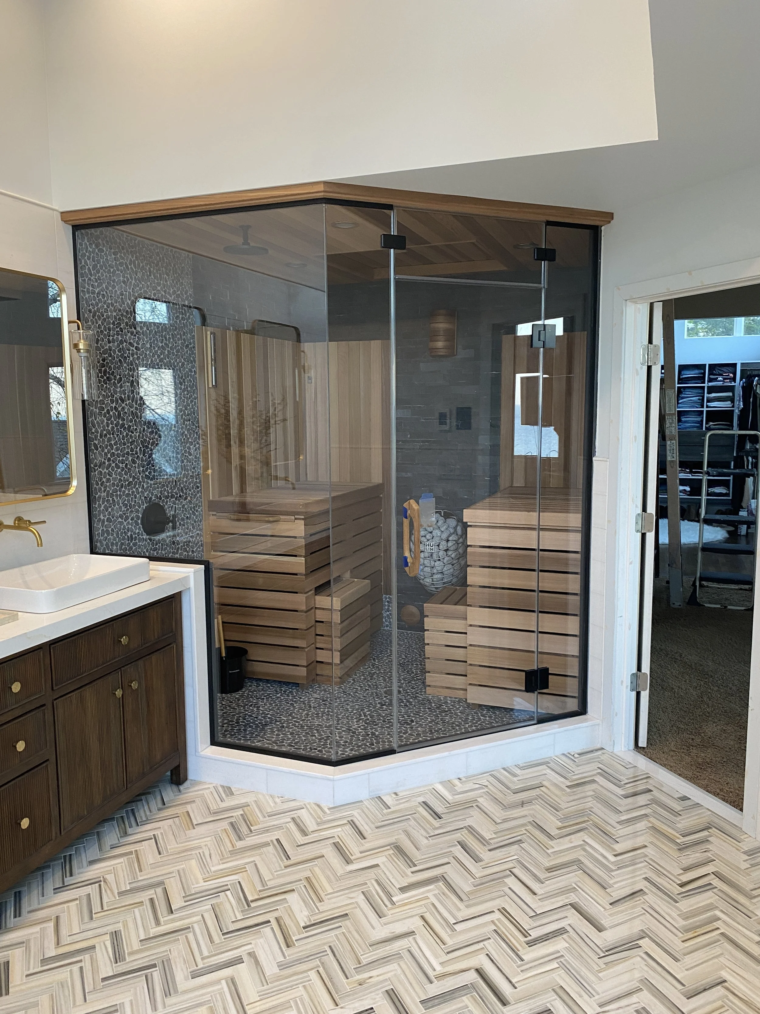 A bathroom with a glass shower enclosure, wooden benches inside, pebble tile on the shower floor, a wood-paneled ceiling, and a patterned tile floor.