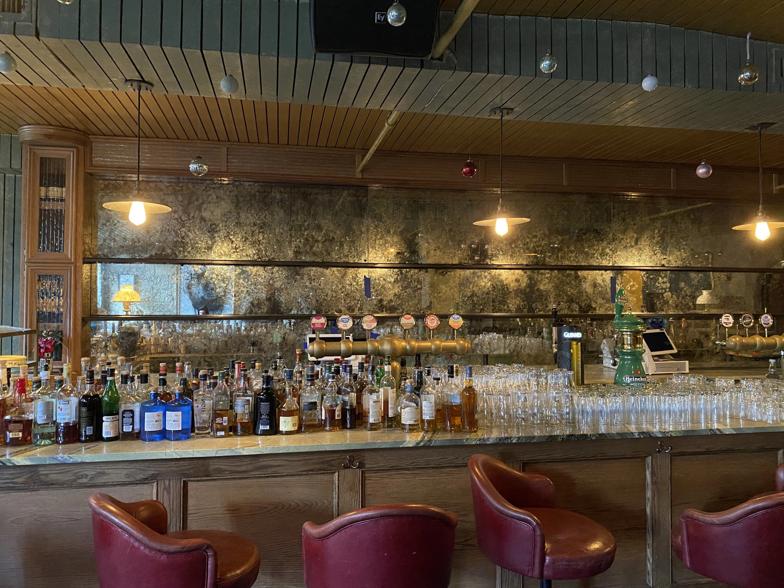 Inside view of a bar with a marble countertop displaying numerous bottles of alcohol, illuminated by warm lighting. The bar has a mirrored backsplash with a textured finish and several hanging pendant lights. There are red leather chairs in the foreground.