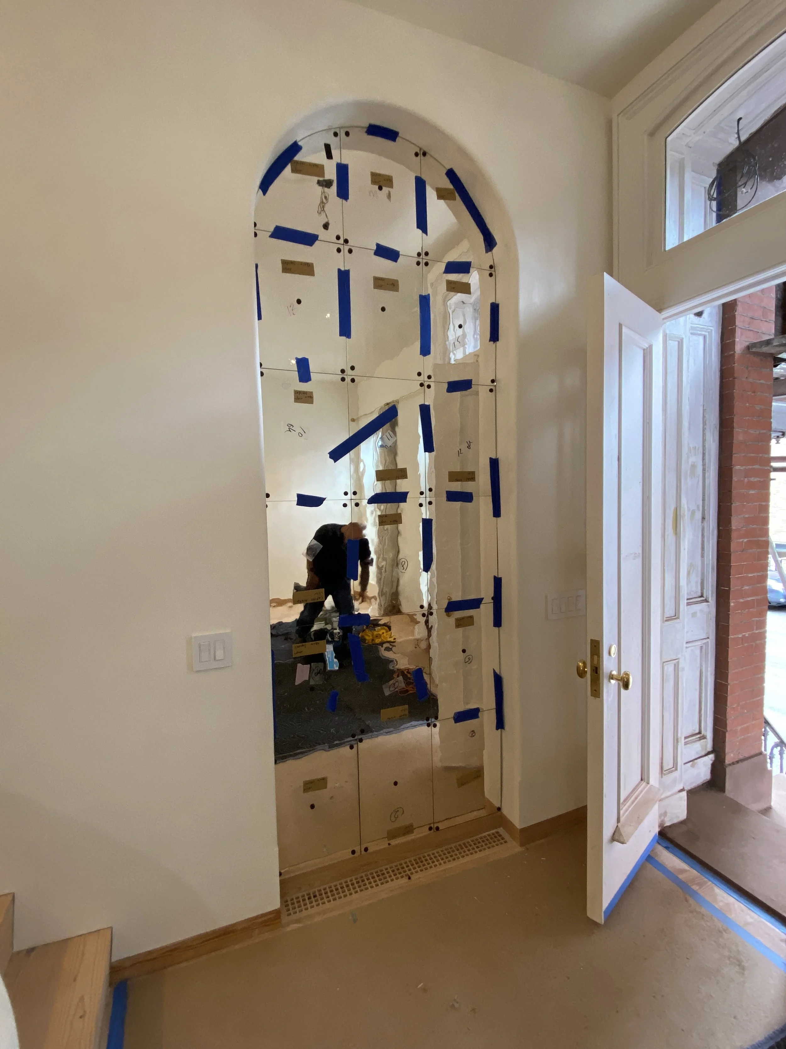 Mirror with materials attached, reflecting the room and a person working nearby, next to a doorway in a house under renovation.