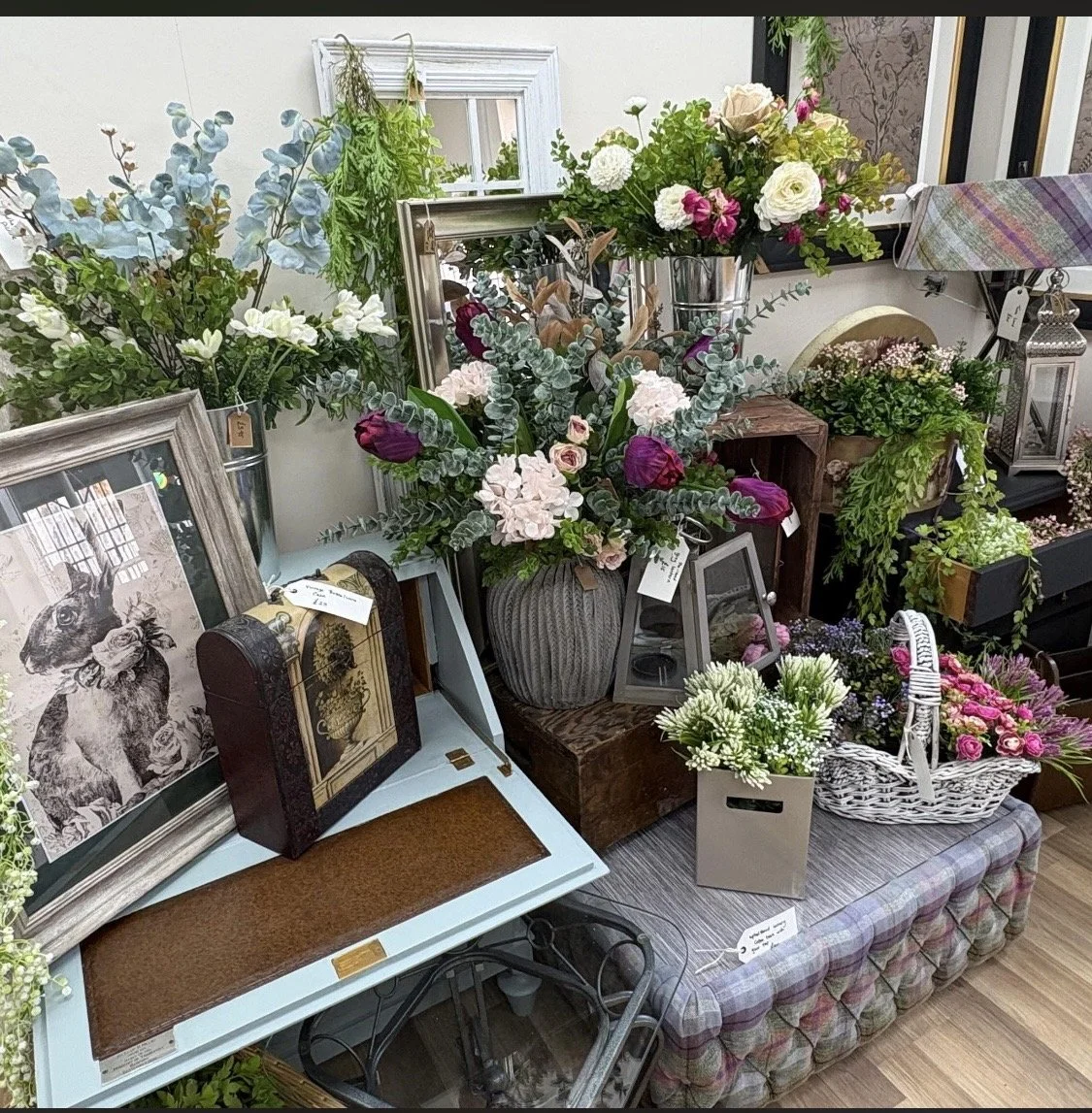 Various flower arrangements and framed artwork displayed on and around a table and shelves in a floral shop or boutique.
