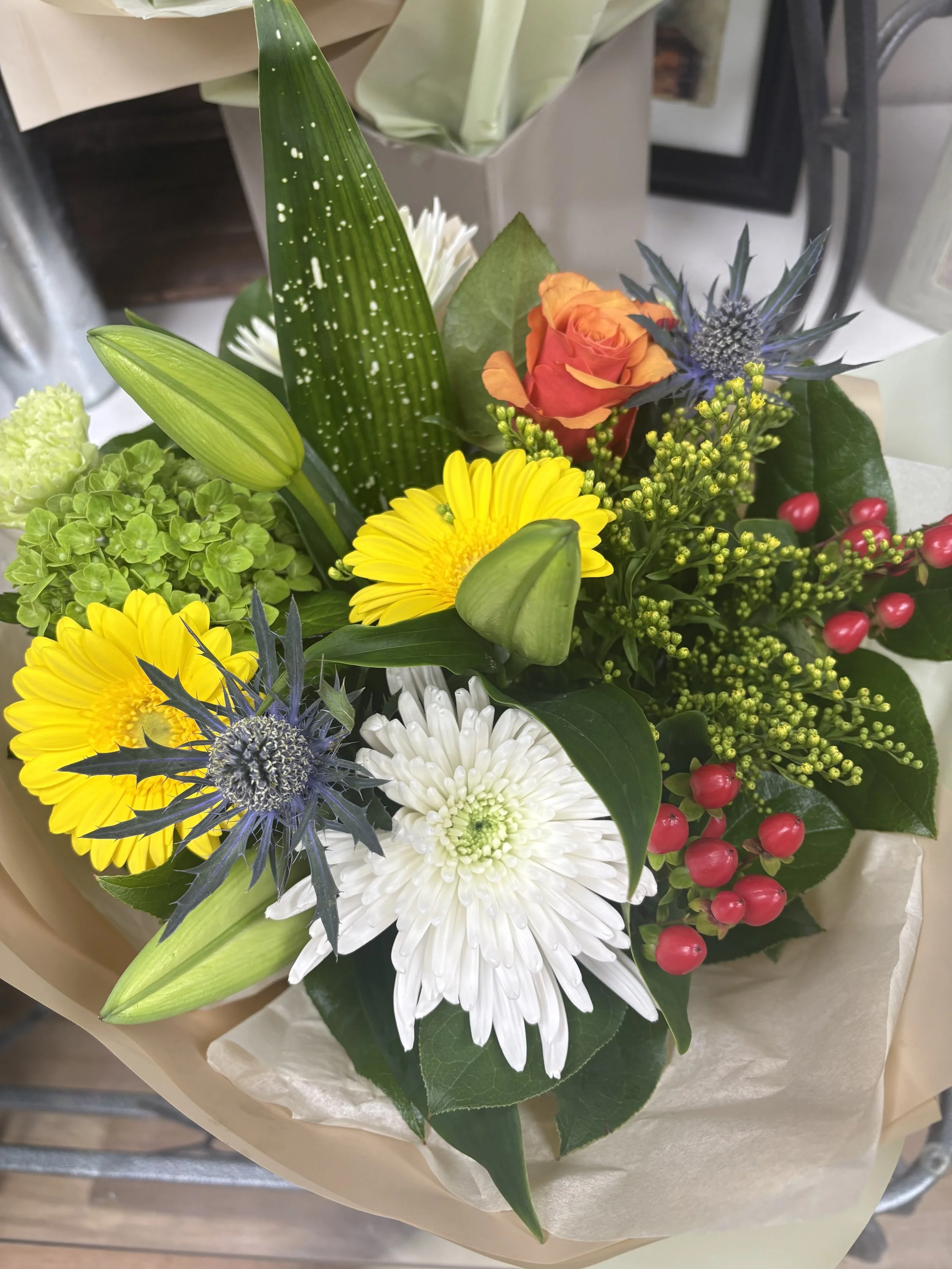 A colorful mixed flower bouquet featuring white, yellow, orange, purple, and red flowers along with green leaves, wrapped in light brown paper.