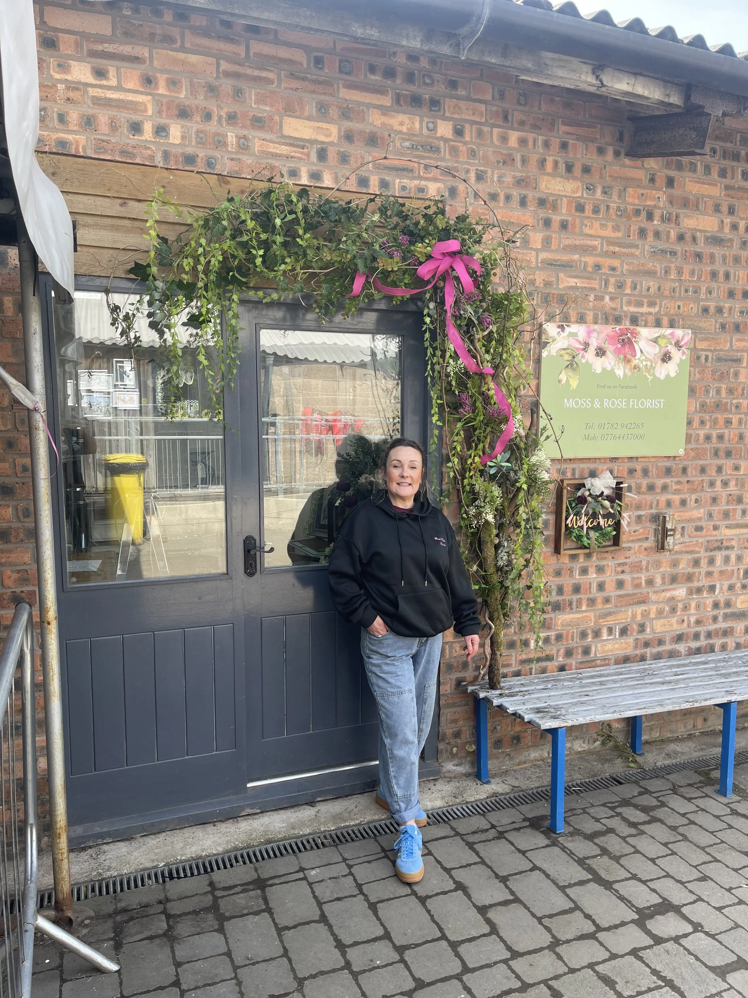 A woman standing outside a brick building in front of a dark gray door decorated with flowers and pink ribbon. She is wearing a black hoodie, light blue jeans, and blue sneakers, with her left hand in her pocket and smiling. There is a green sign on the wall for Moss & Rose Florist and a wooden bench beneath it.