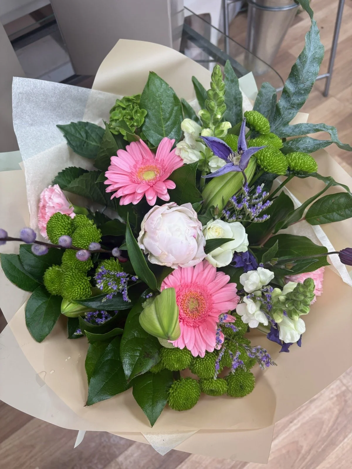 A colorful bouquet of fresh flowers including pink gerbera daisies, white peonies, purple clematis, green chrysanthemums, and various green leaves, wrapped in light-colored paper.