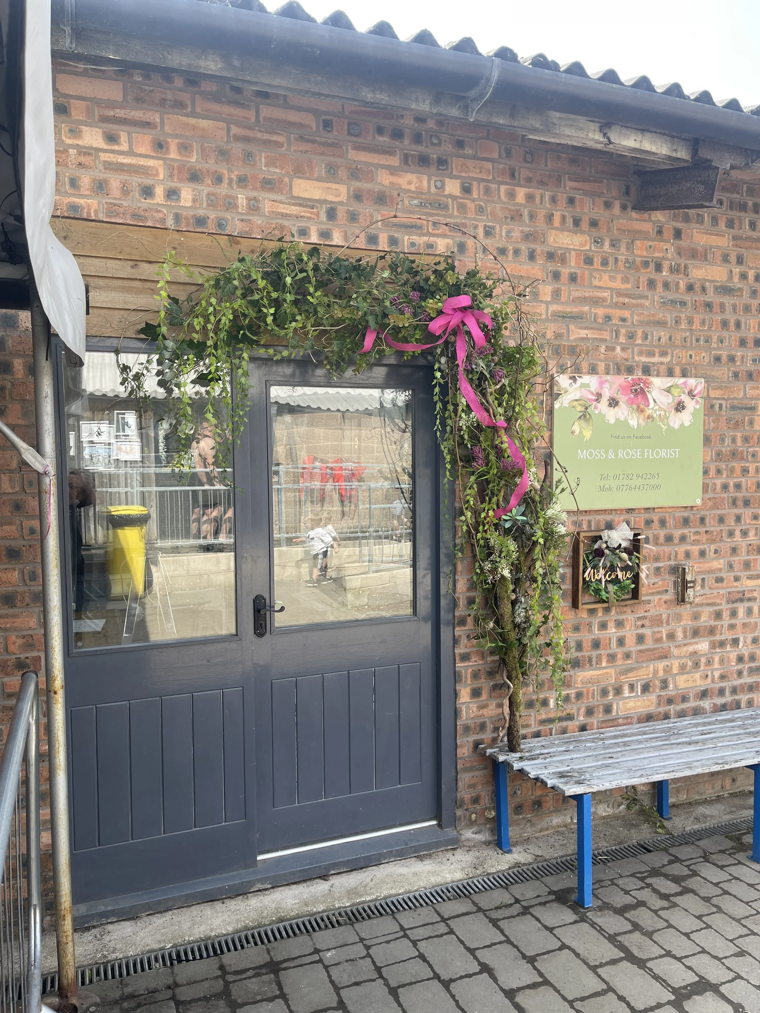 The image shows a grey door with a window in the center, on a brick wall building. A pink ribbon is tied in a bow on a decorative arrangement of greenery and flowers above the door. To the right, there is a green sign that reads 'Moss & Rose Florist' with contact information. A small wooden 'Welcome' sign with floral decorations is mounted on the wall below the sign. A blue bench is positioned on the right side next to a small tree. Part of a metal scaffolding and a yellow trash bin are visible on the left side. The ground is paved with cobblestones, and a drainage grate runs along the front of the door.