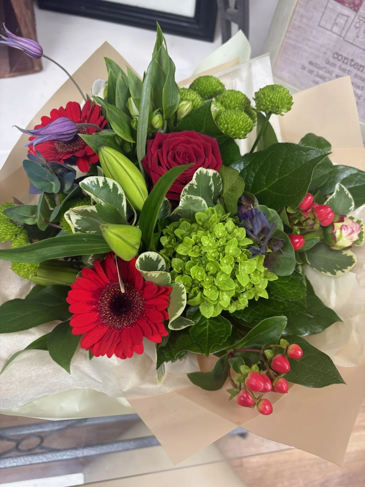 A colorful bouquet of flowers including red gerbera daisies, a red rose, green hydrangea, purple flowers, green berries, and various greenery, wrapped in white and beige paper.