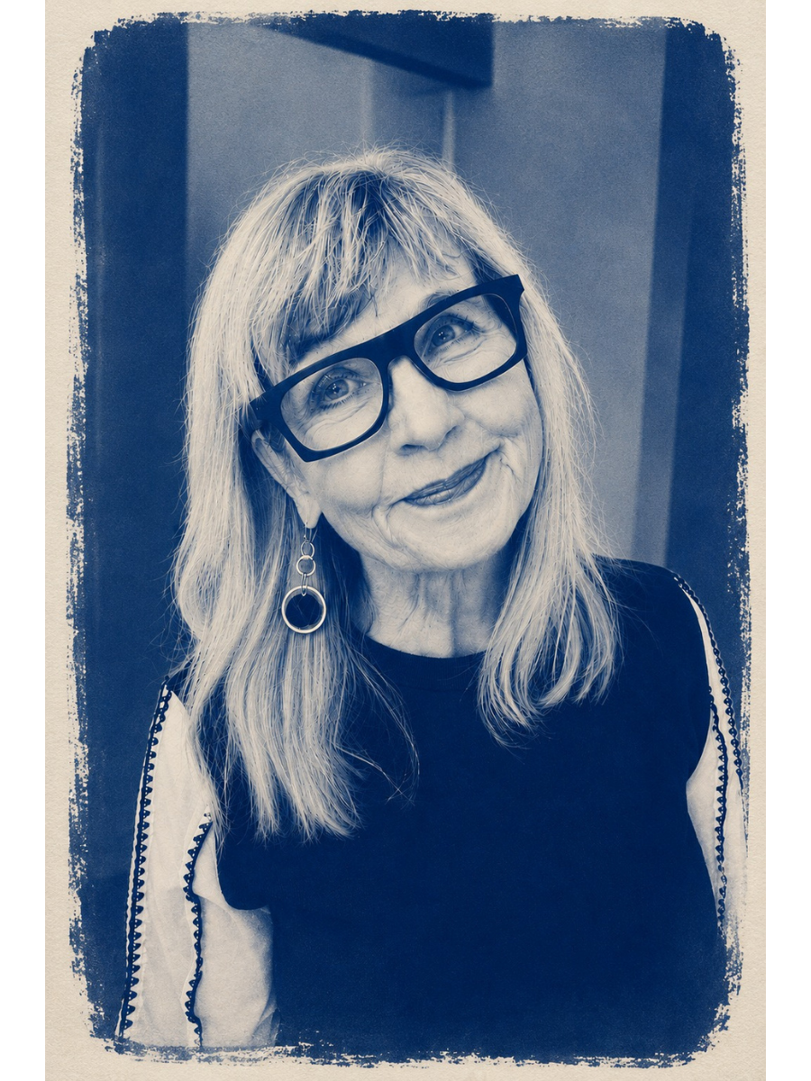 A smiling woman with long blonde hair, wearing large black glasses and hoop earrings, posing indoors against a neutral background.
